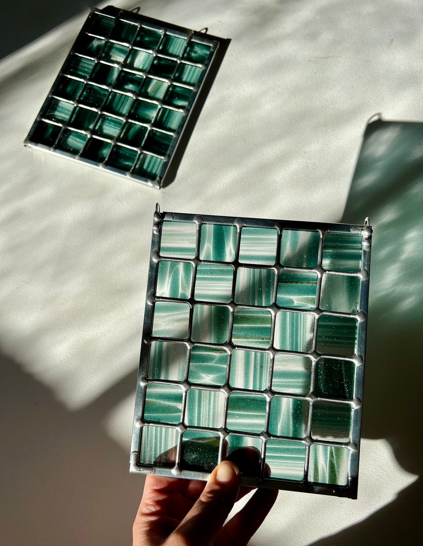 hand holding a stained glass suncatcher depicting streaky and mottled sparkly clear and teal glass arranged in a grid pattern, held above a white surface with sunlight casting a partial refraction, with a second similar suncatcher in the background