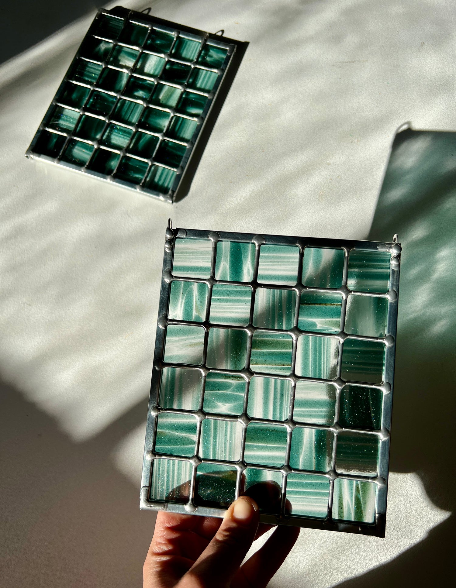 hand holding a stained glass suncatcher depicting streaky and mottled sparkly clear and teal glass arranged in a grid pattern, held above a white surface with sunlight casting a partial refraction, with a second similar suncatcher in the background