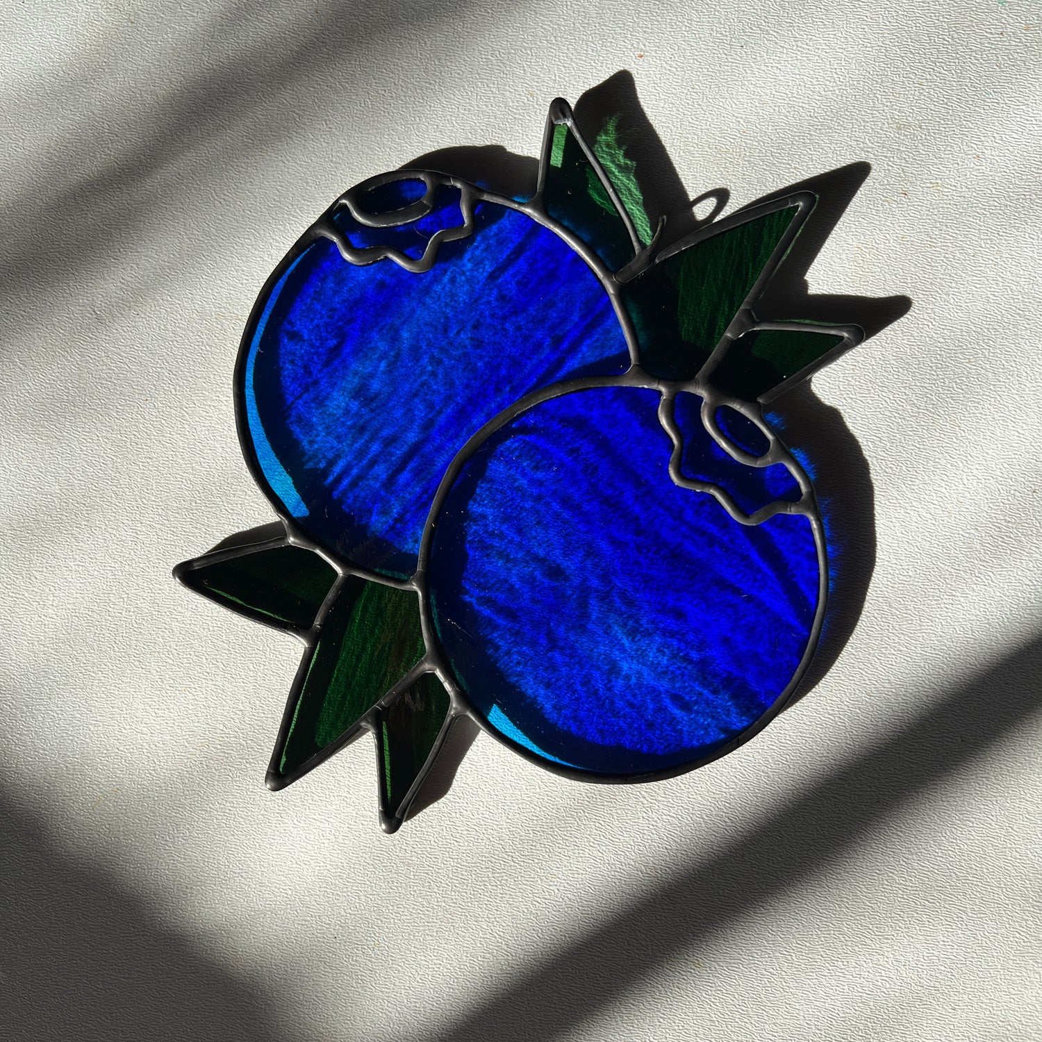 a wispy blue stained glass suncatcher depicting a small bunch of blueberries with leaves resting on a white surface