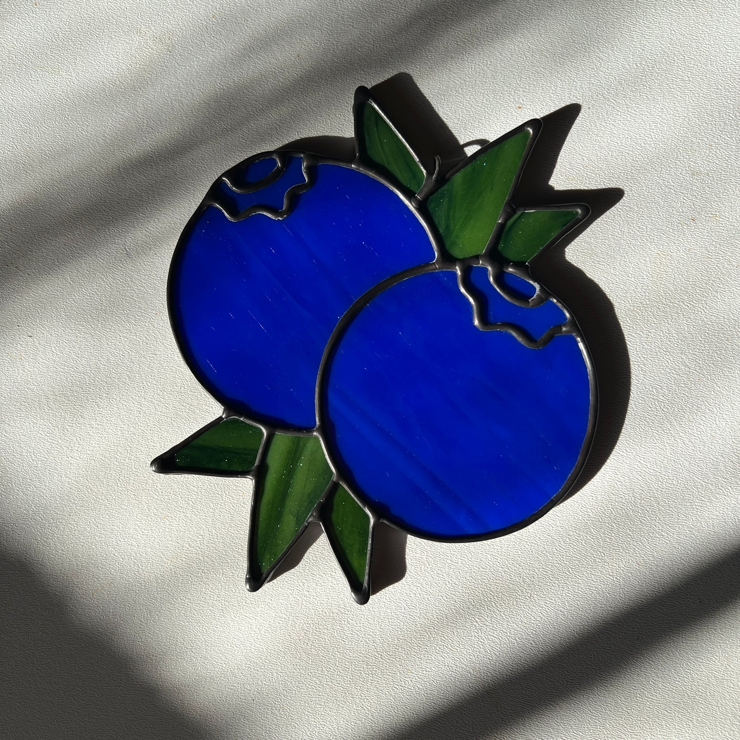 stained glass suncatcher depicting a small bunch of blueberries with leaves resting on a white surface