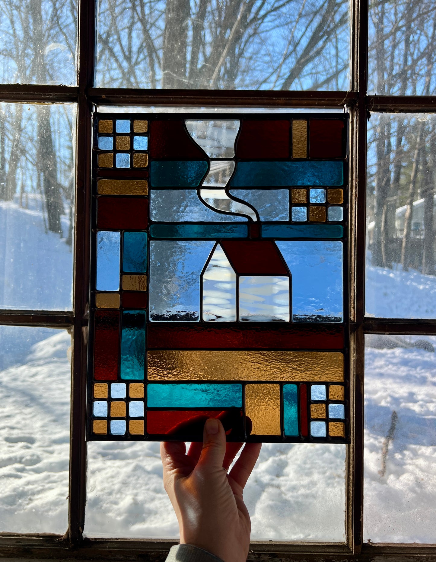 Stained glass textile inspired panel depicting a home in the center in shades of teal, brown, and blue, held by a hand against a large window with a snowy landscape outside