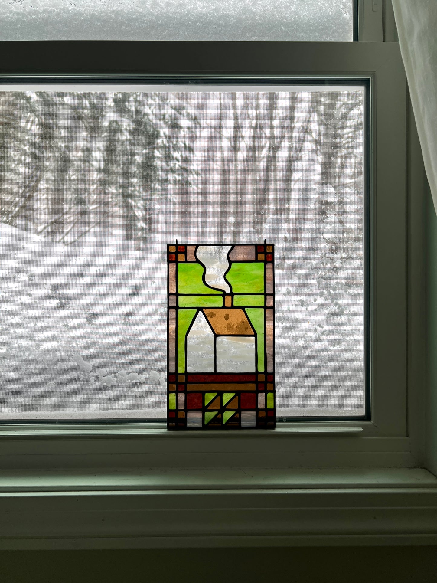 Stained glass textile inspired panel depicting a home in the center in shades of green, pink, white, and brown, resting in a window with a snowy scene outside.