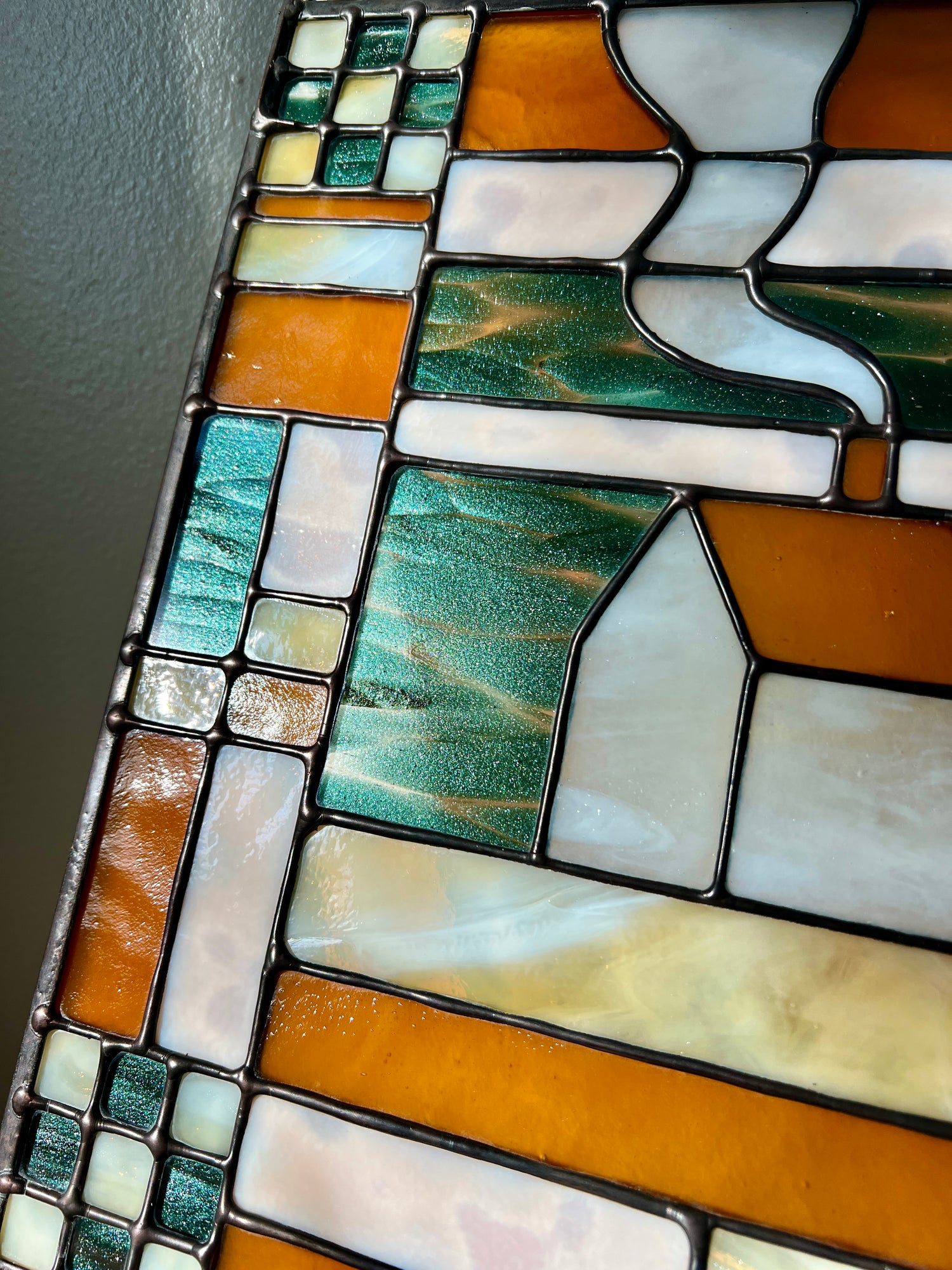 Close up of a stained glass textile inspired panel depicting a home in the center in shades of teal, amber, white, and beige, highlighting smooth solder lines and unique glitter effect of the teal glass