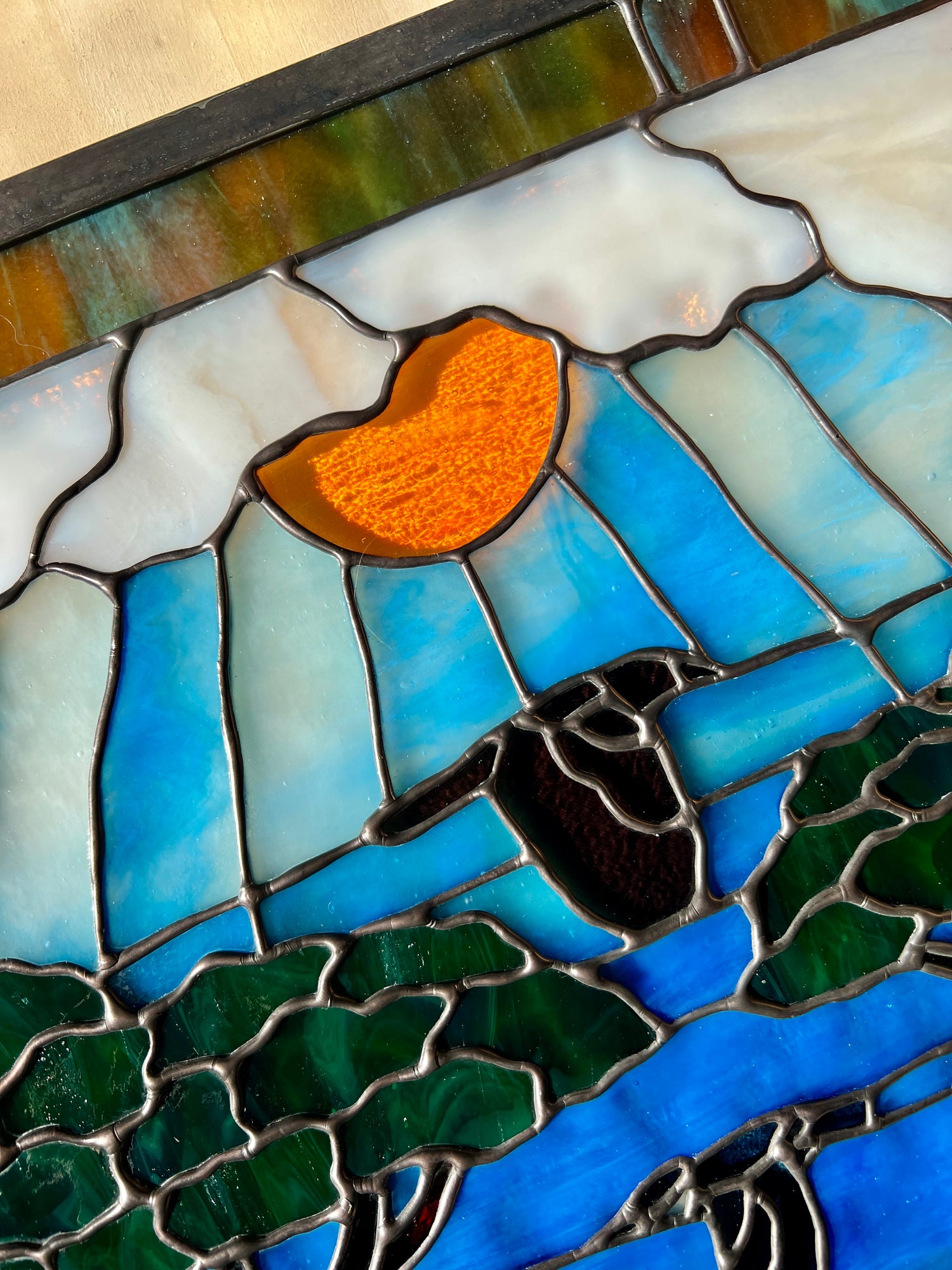 close up of a large stained glass panel depicting mossy, rocky cliffs and trees, with a deer, a kingfisher, and a raven standing amongst a river, greenery, and bright blue skies with some clouds and a bright yellow orange sun.