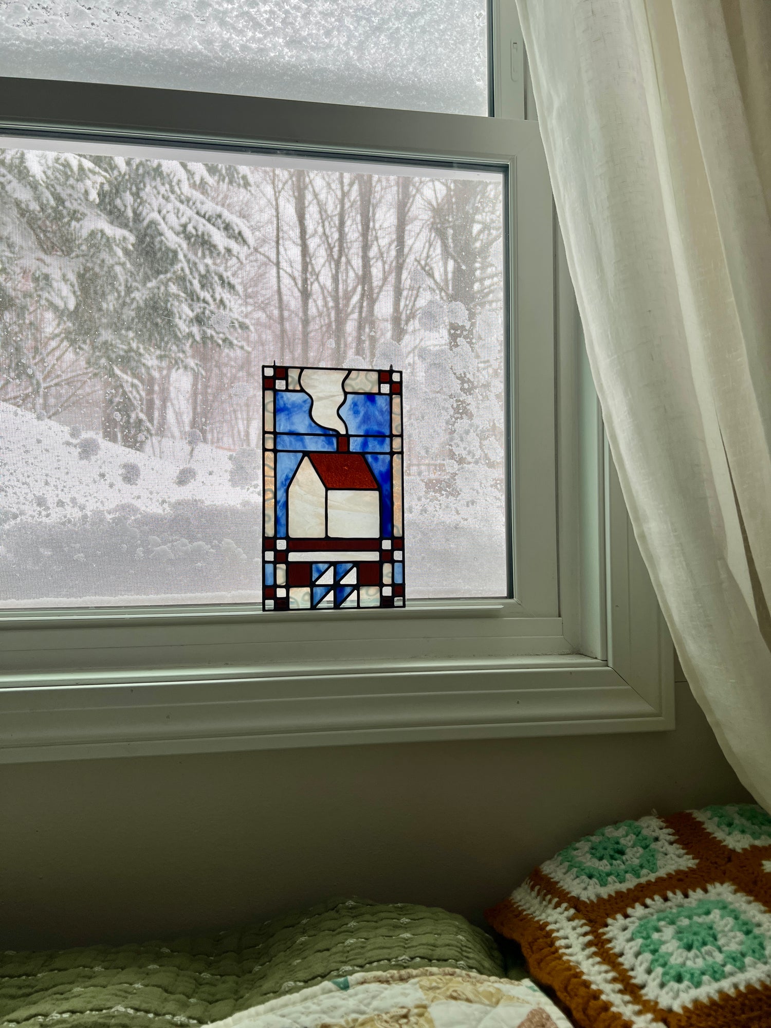 Stained glass textile inspired panel depicting a home in the center in shades of blue, brown, and white, resting in a window with a snowy landscape outside and cozy blankets in the foreground