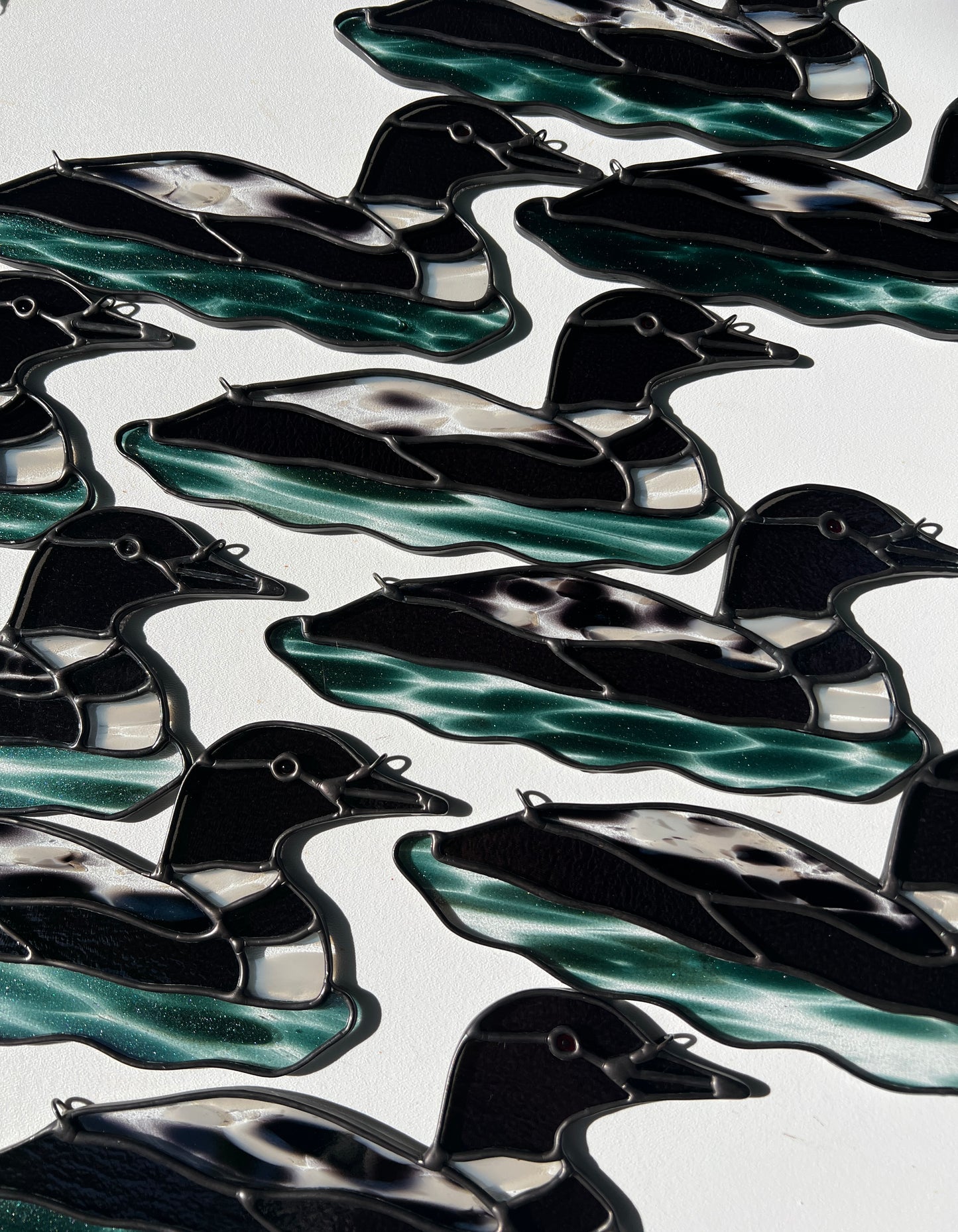 ten stained glass loons arranged on a white surface with bright sunlight cast across, photographed at an angle