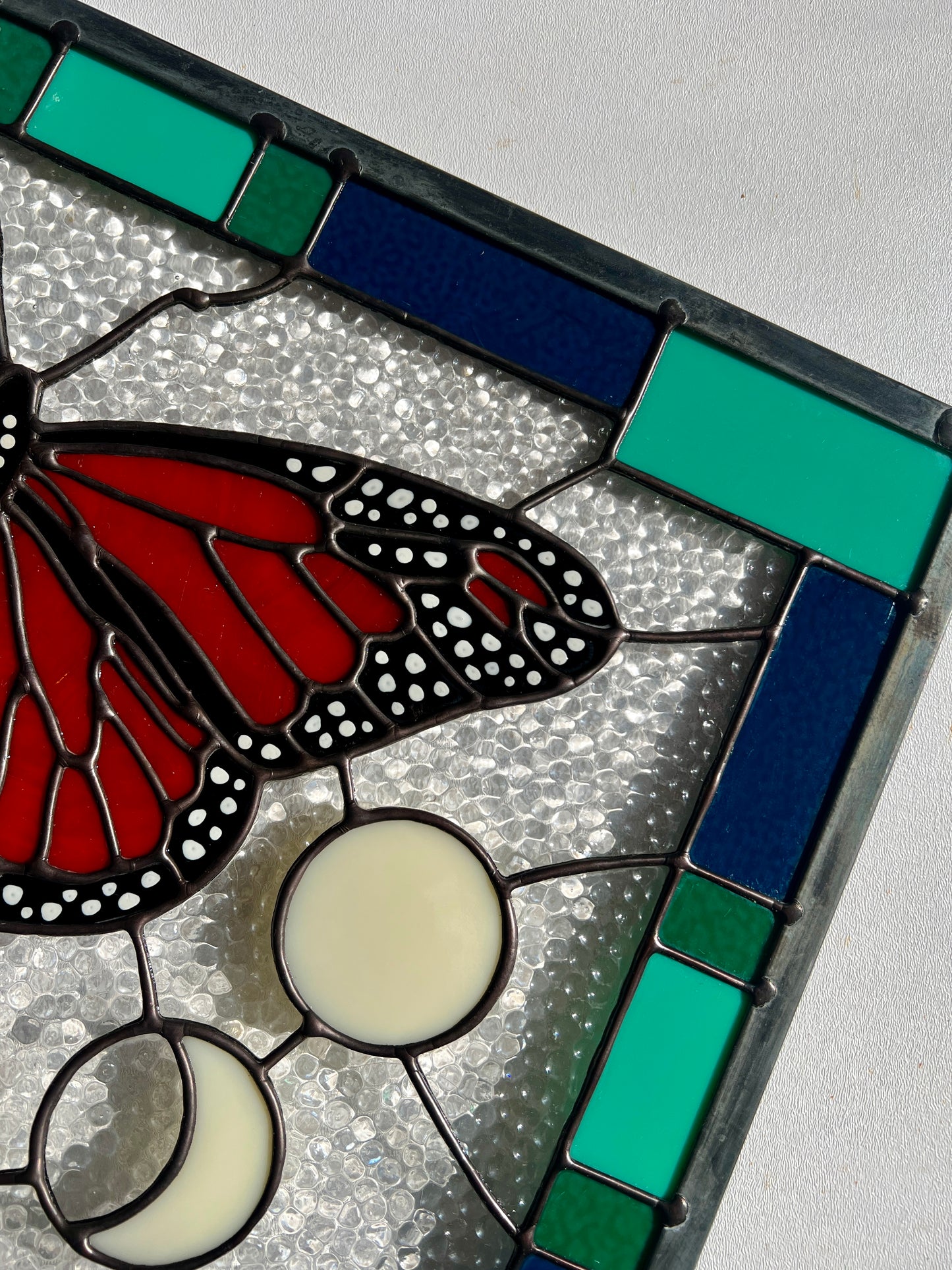 Close up of the hand painted details of a large stained glass panel with a thick blue and green geometric border and a monarch butterfly and moon phases in the center, with textured clear glass in the background.
