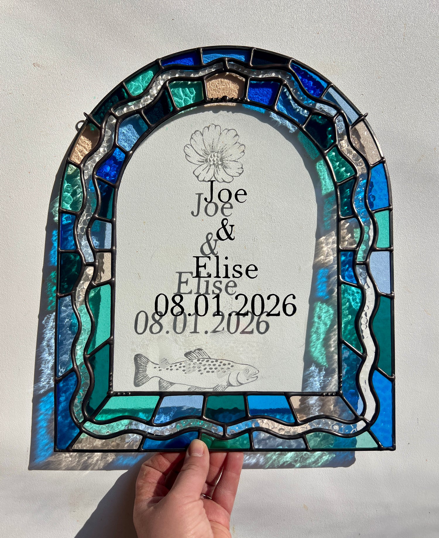 Stained glass panel in the shape of an arch with a multicolor blue, teal, turquoise, and pink border. Within the border is squiggle detail in clear textured glass. The center of the arch is clear uncolored glass, with personalized text and hand etched details of a cosmos flower and trout. The piece is held by a hand against a white wall, with sunlight casting refractions and shadows of the text, colored glass, and etchings.