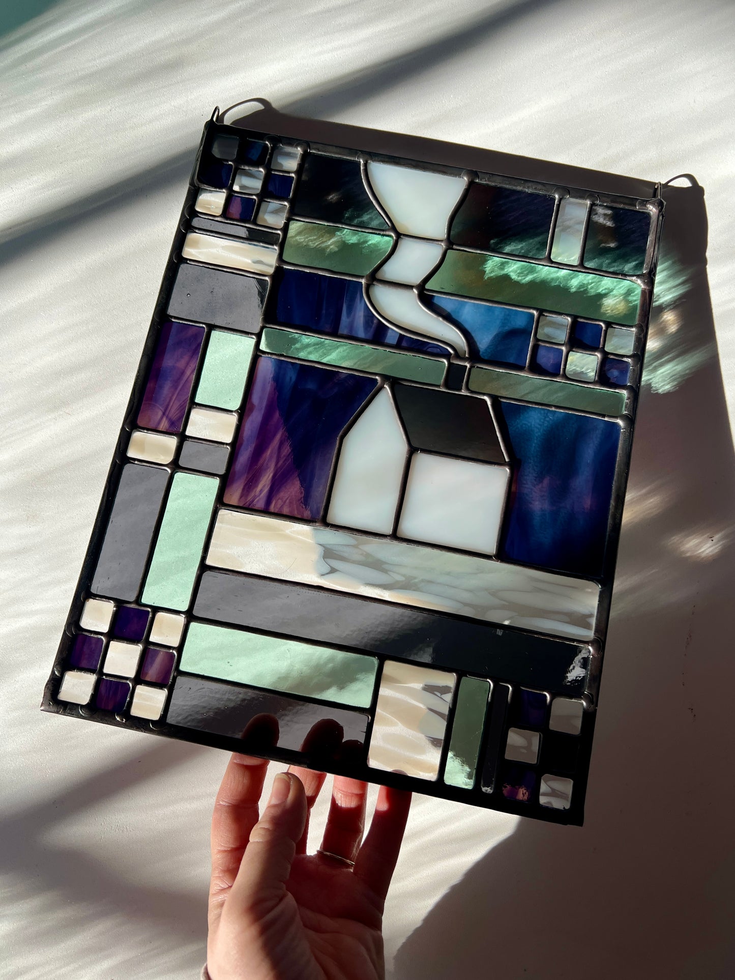Stained glass panel with geometric textile inspired design with a cozy home depicted in the center, held by a hand against a white background with sun and shadows flowing across it.