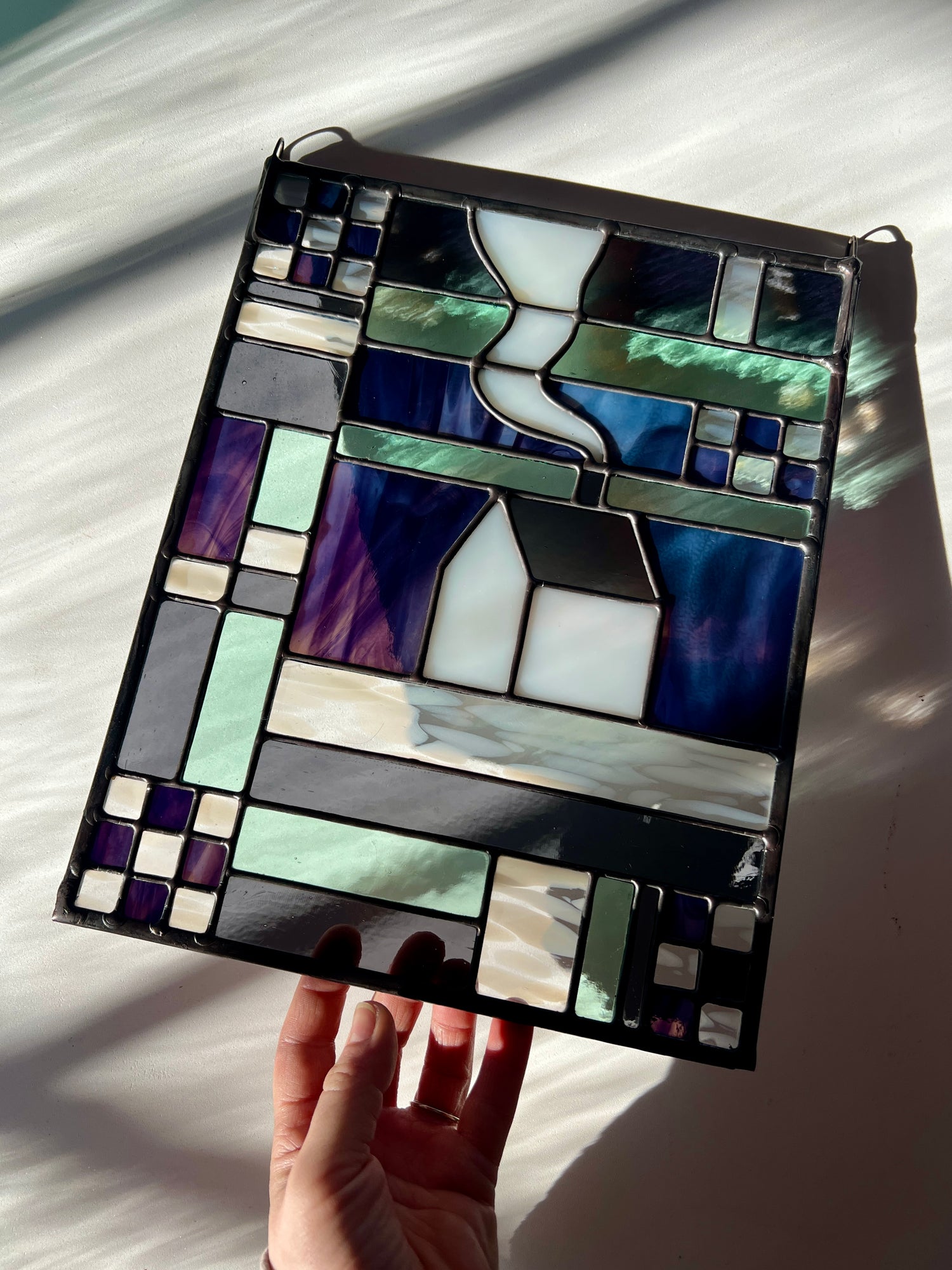 Stained glass panel with geometric textile inspired design with a cozy home depicted in the center, held by a hand against a white background with sun and shadows flowing across it.