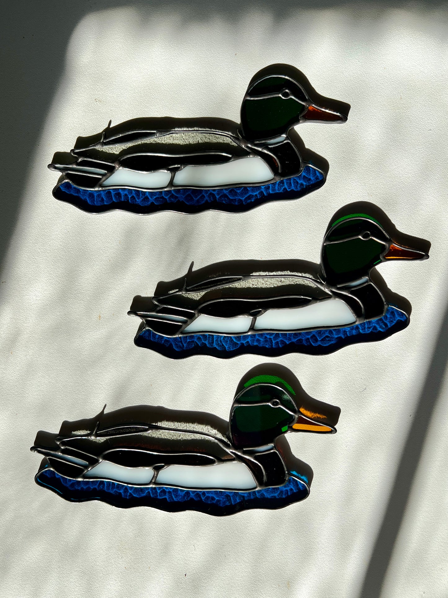 three stained glass suncatchers depicting male mallards resting on a white surface in bright sunlight
