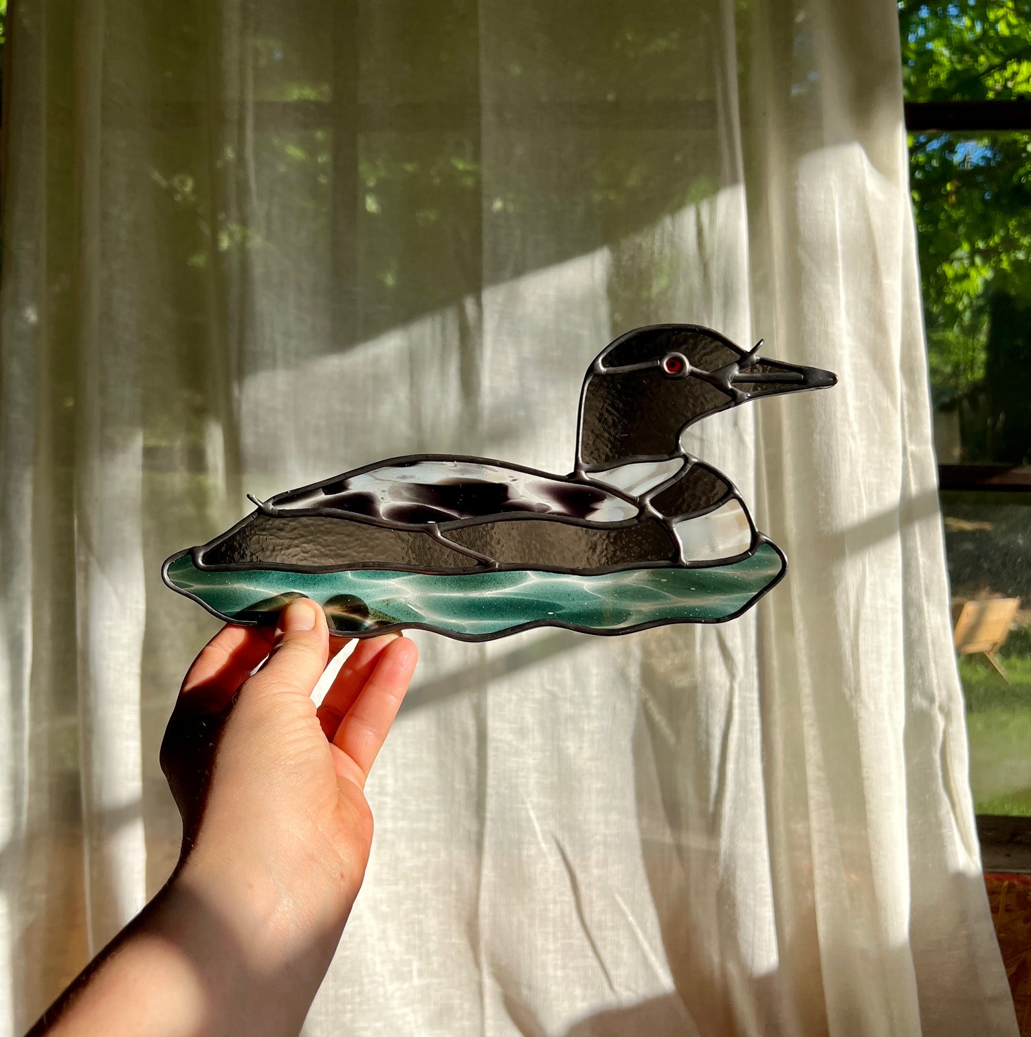 hand holding a stained glass loon in front of a large window with a sheer curtain