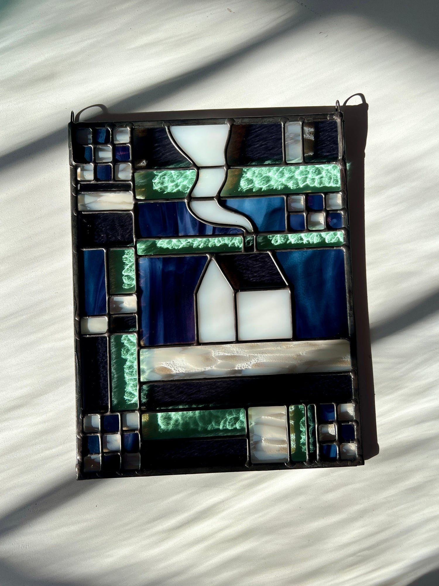 Stained glass panel with geometric textile inspired design with a cozy home depicted in the center, laying on a white black ground with bright sunlight across it