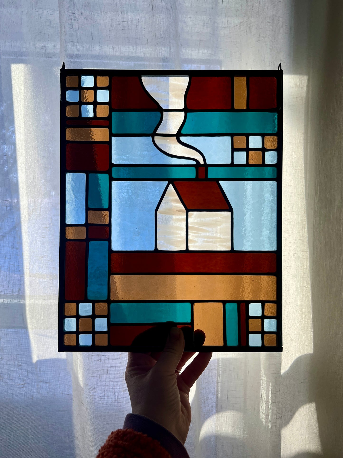 Stained glass textile inspired panel depicting a home in the center in shades of teal, brown, and blue, held by a hand against a sheer white curtain that is gently backlit by morning sunlight