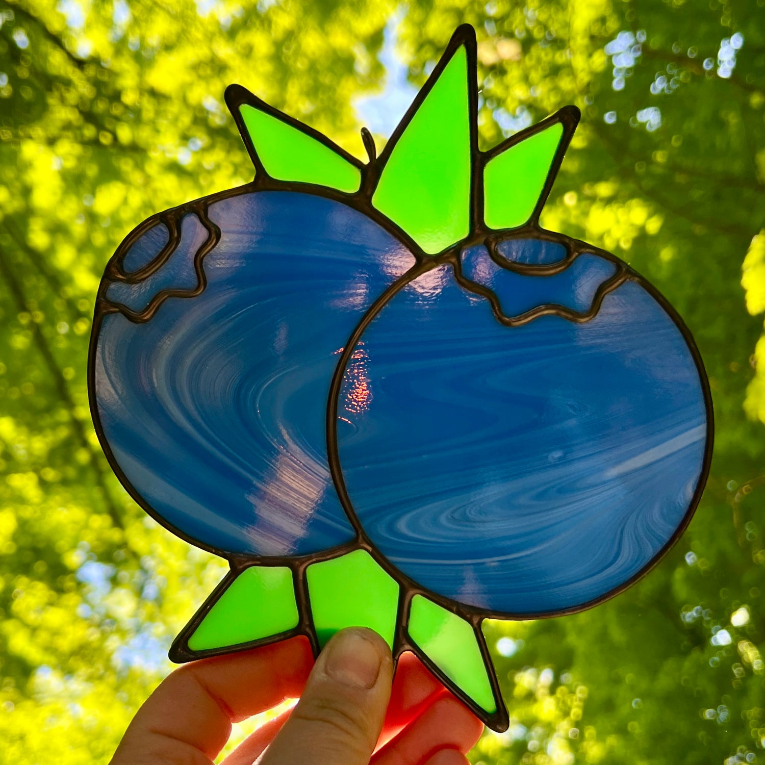hand holding a streaky stained glass suncatcher depicting a small bunch of blueberries with leaves, held against sunlight with bright green leaves and trees in the background
