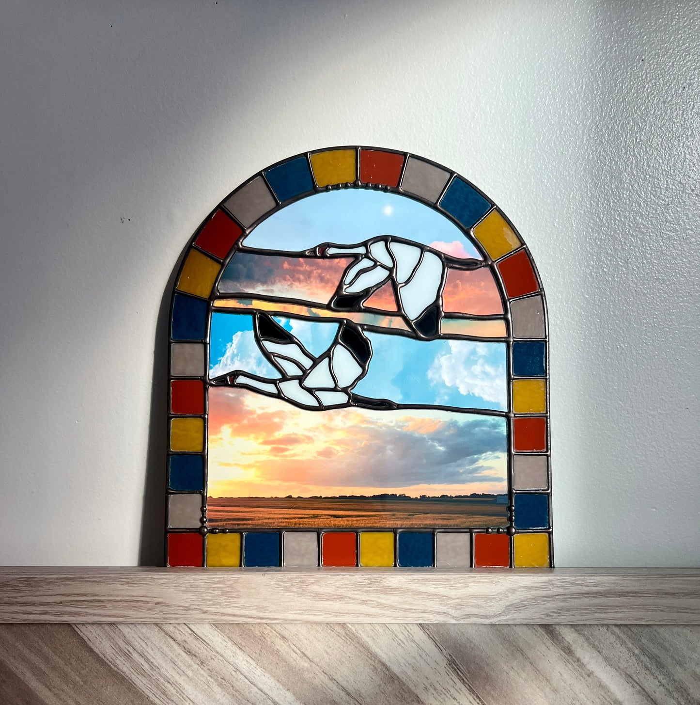 Stained glass panel with a colorful border featuring two birds and a sunset scene on a white wall.