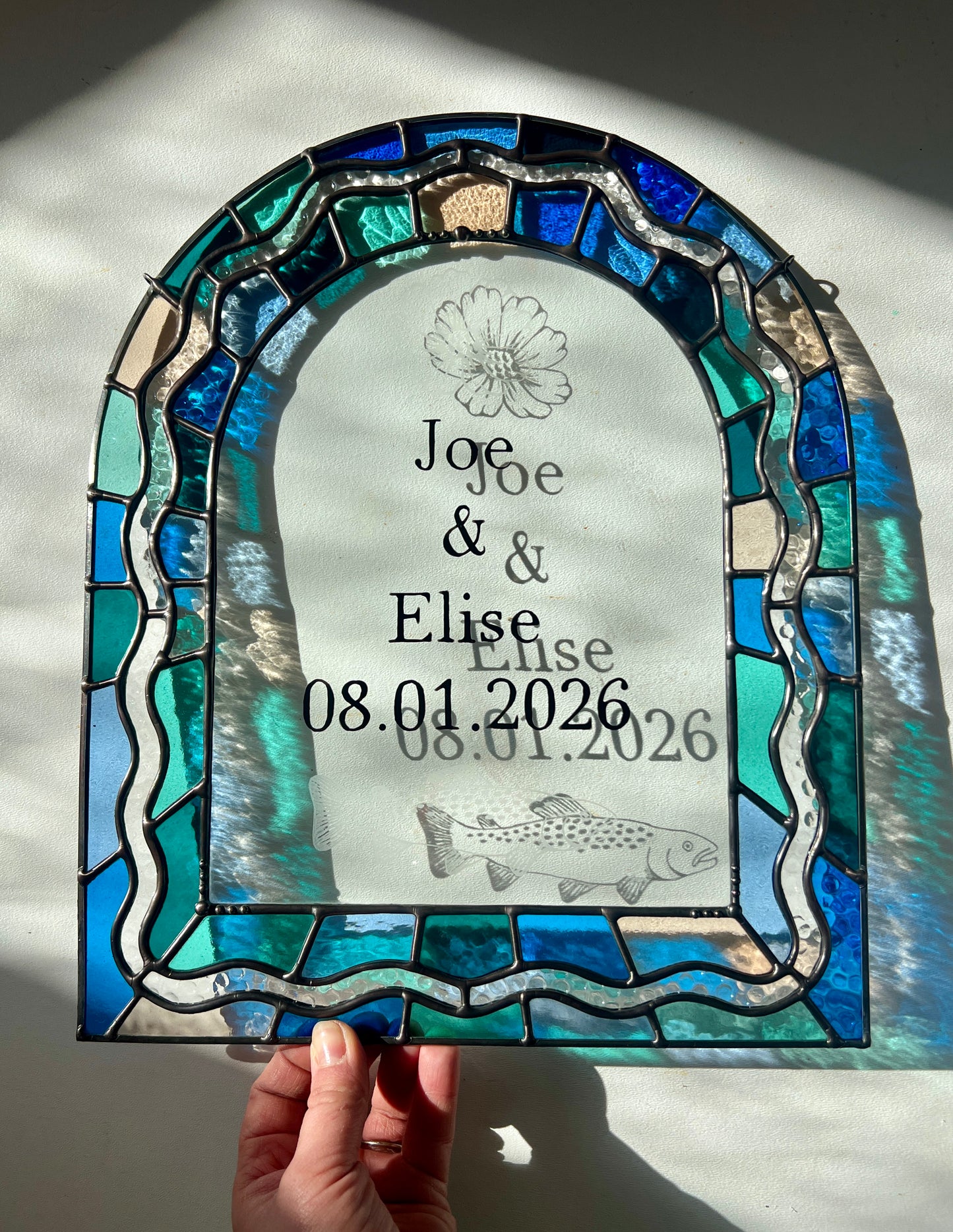 Stained glass panel in the shape of an arch with a multicolor blue, teal, turquoise, and pink border. Within the border is squiggle detail in clear textured glass. The center of the arch is clear uncolored glass, with personalized text and hand etched details of a cosmos flower and trout. The piece is held by a hand against a white wall, with sunlight casting refractions and shadows of the text, colored glass, and etchings.