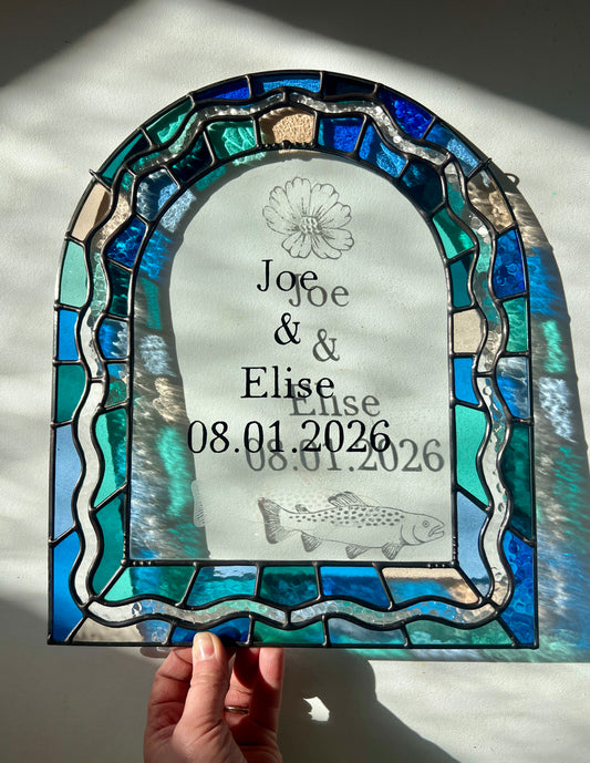 Stained glass panel in the shape of an arch with a multicolor blue, teal, turquoise, and pink border. Within the border is squiggle detail in clear textured glass. The center of the arch is clear uncolored glass, with personalized text and hand etched details of a cosmos flower and trout. The piece is held by a hand against a white wall, with sunlight casting refractions and shadows of the text, colored glass, and etchings.