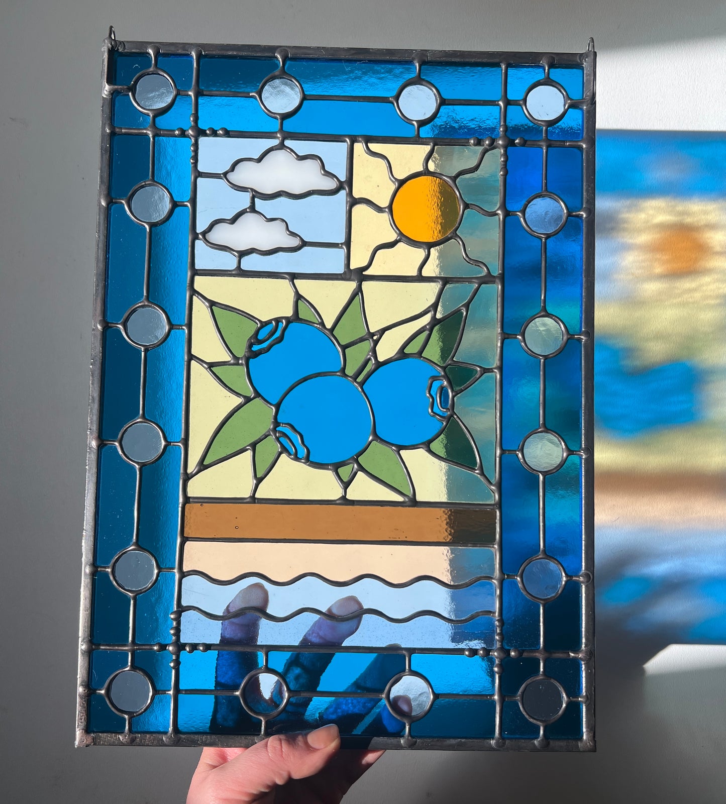 stained glass panel with a simple compartment style design featuring clouds, sun, water, and a small bunch of blueberries in the middle with a blue border