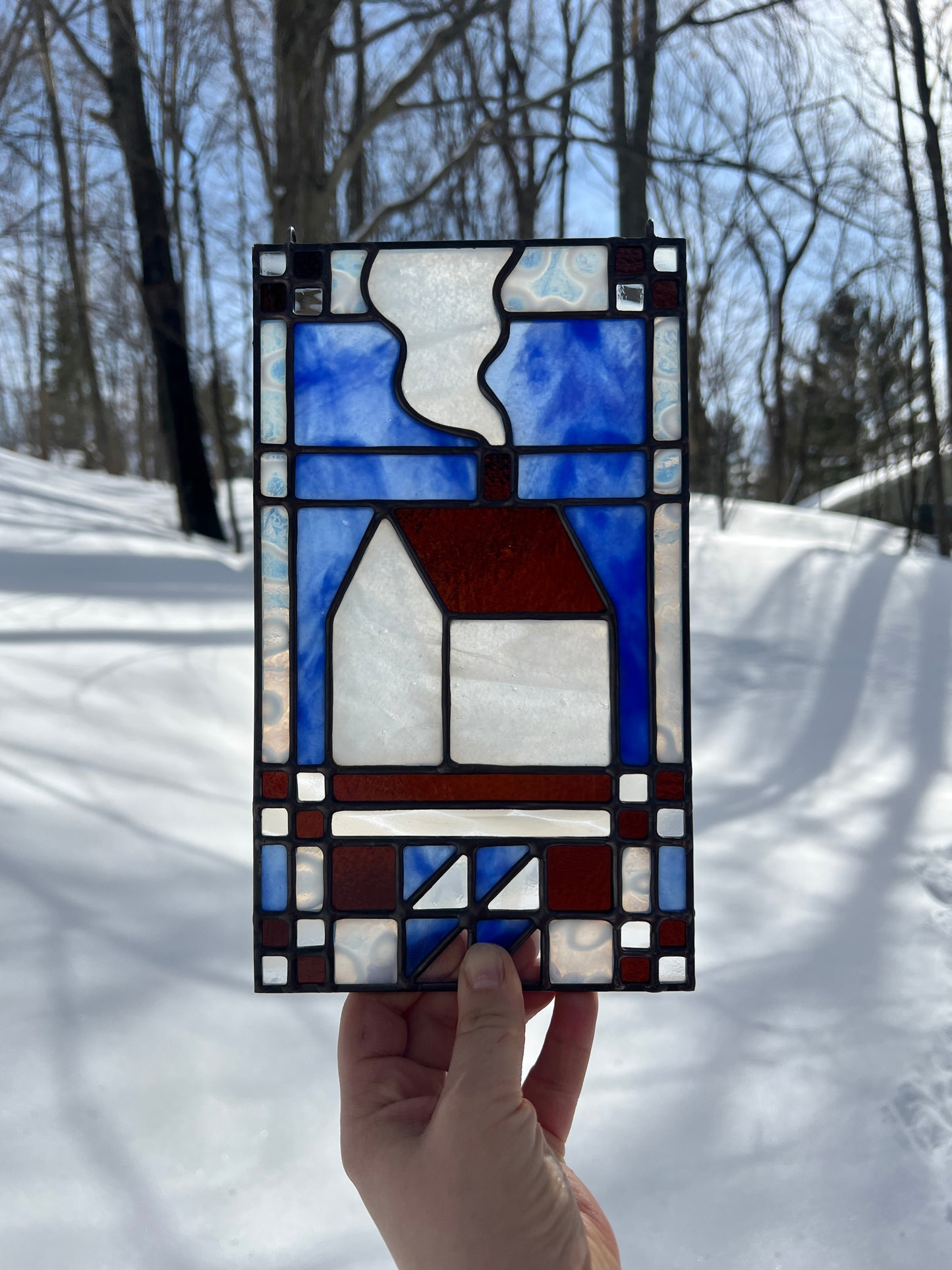 Stained glass textile inspired panel depicting a home in the center in shades of blue, brown, and white, held by a hand in front of a snow covered landscape
