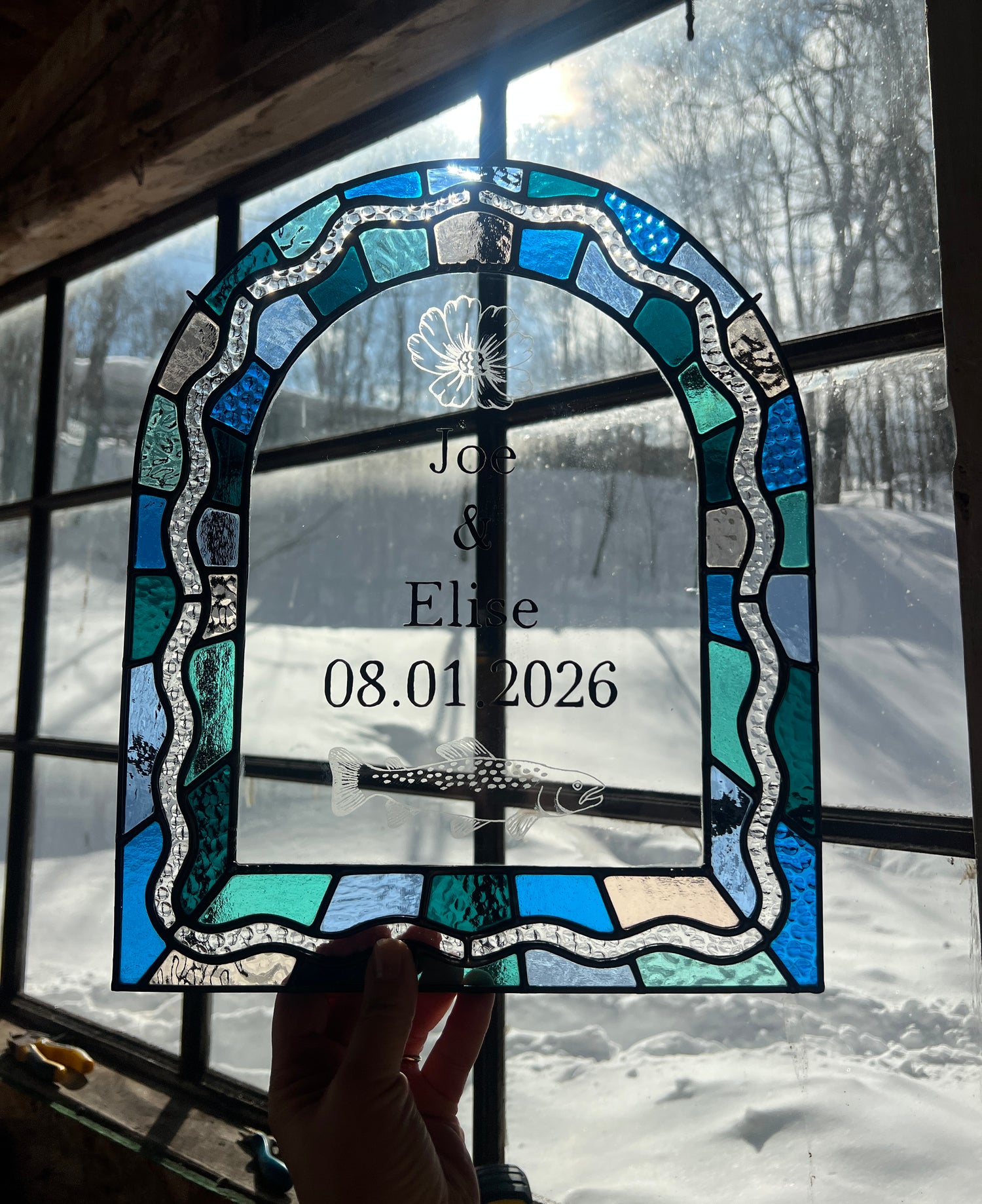 Stained glass panel in the shape of an arch with a multicolor blue, teal, turquoise, and pink border. Within the border is squiggle detail in clear textured glass. The center of the arch is clear uncolored glass, with personalized text and hand etched details of a cosmos flower and trout. The piece is held by a hand and held up to a window with snow outside.