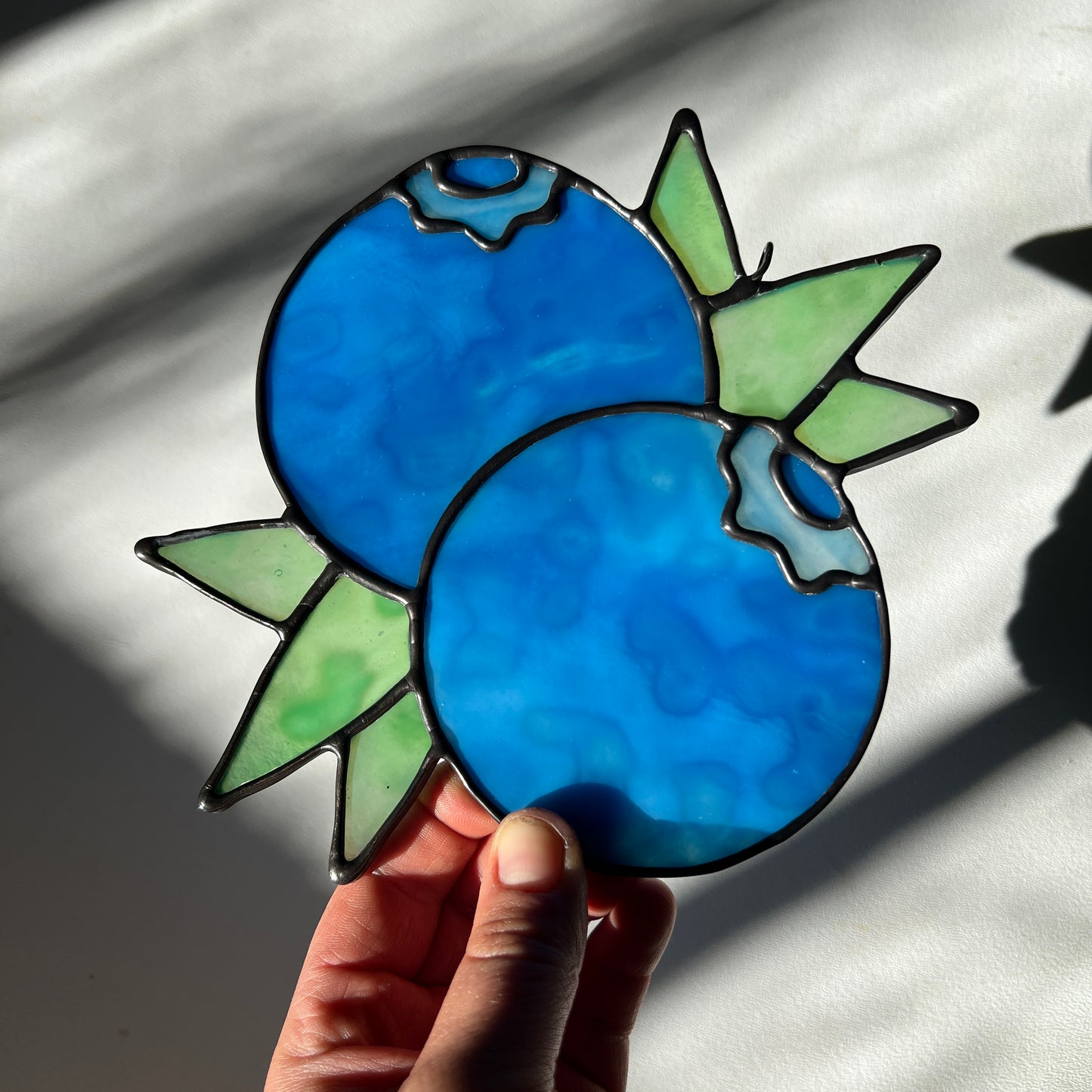 hand holding a mottled blue stained glass suncatcher depicting a small bunch of blueberries with leaves