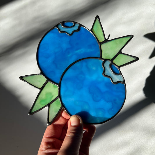 hand holding a mottled blue stained glass suncatcher depicting a small bunch of blueberries with leaves