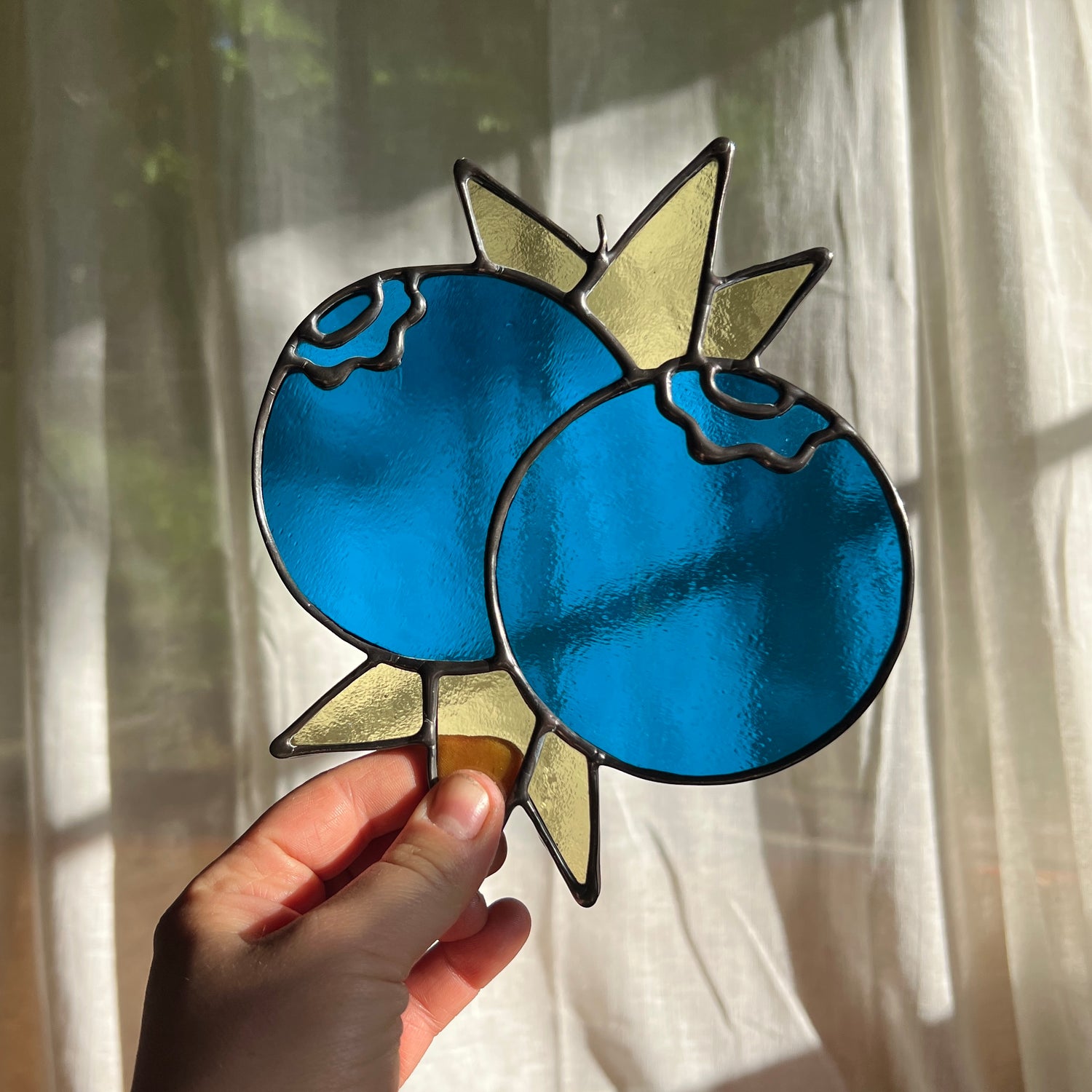 hand holding a stained glass suncatcher depicting a small bunch of blueberries with leaves in front of a window