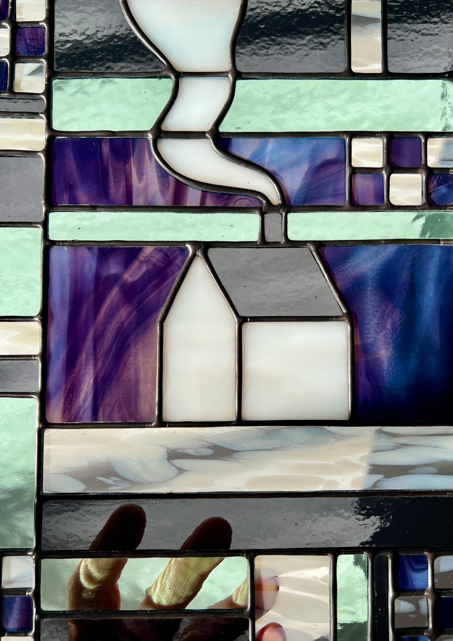 Close up of a stained glass geometric textile inspired panel depicting a cozy home in the center, with soft backlighting showcasing vibrant shades of purple, soft shades of gray, and pastel shades of sea foam.