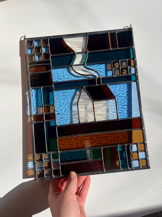 Stained glass textile inspired panel depicting a home in the center in shades of teal, brown, and blue, held by a hand against a white background