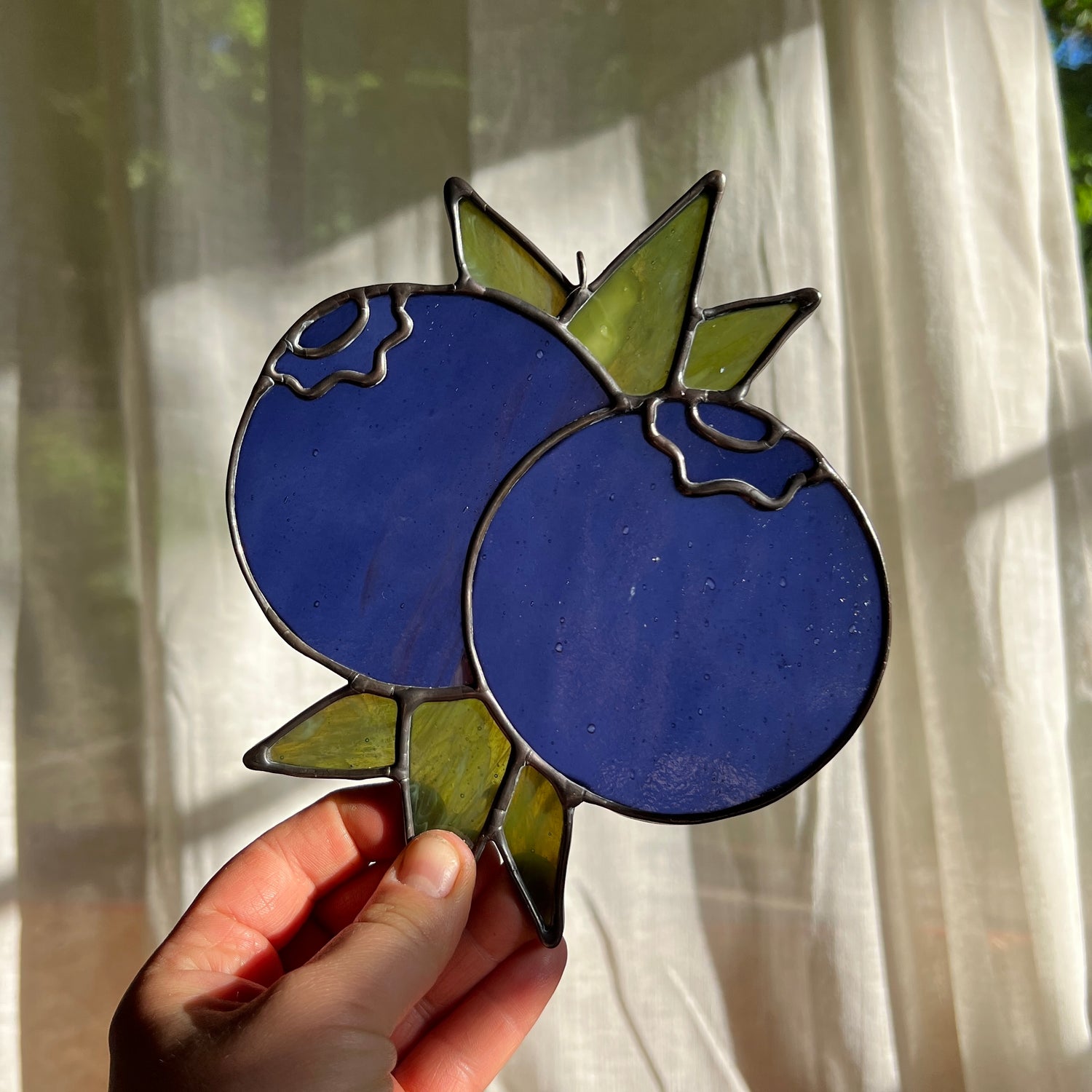 hand holding a blue stained glass suncatcher depicting a small bunch of blueberries with leaves in front of a window