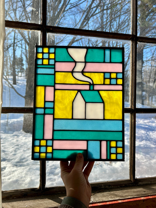 Stained glass textile inspired panel depicting a home in the center in shades of teal, pink, white, blue, and yellow, held by a hand in front of a window with a snowy scene outside.