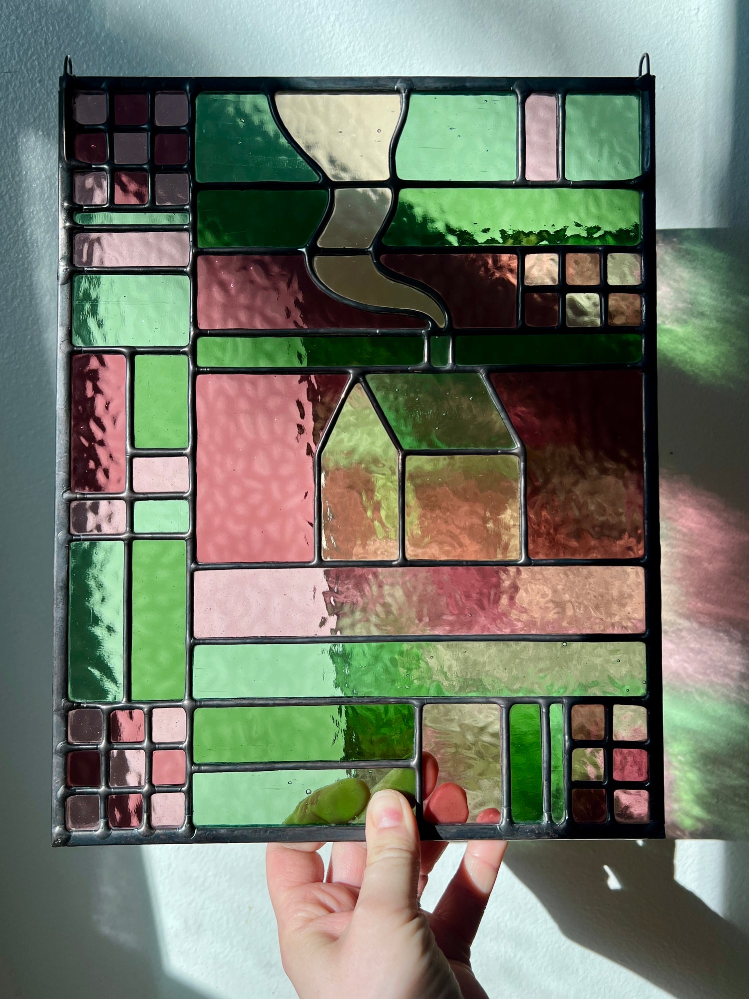 Stained glass panel with geometric textile inspired design with a cozy home depicted in the center, held by a hand up to a white back ground, with sunlight casting pink and green refractions from the glass