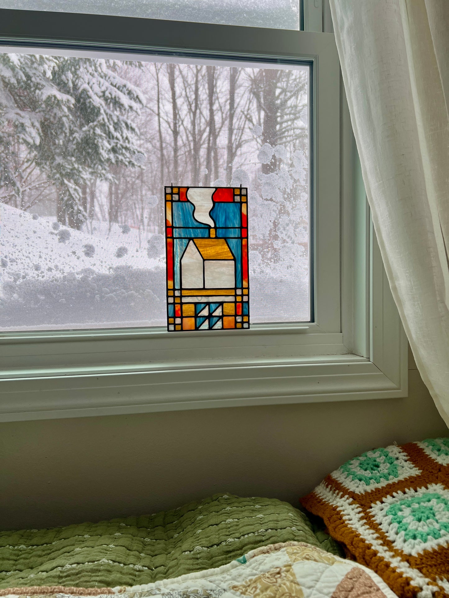 Stained glass textile inspired panel depicting a home in the center in shades of red, blue, white, and amber, resting in a window with a snowy scene outside and cozy blankets in the foreground