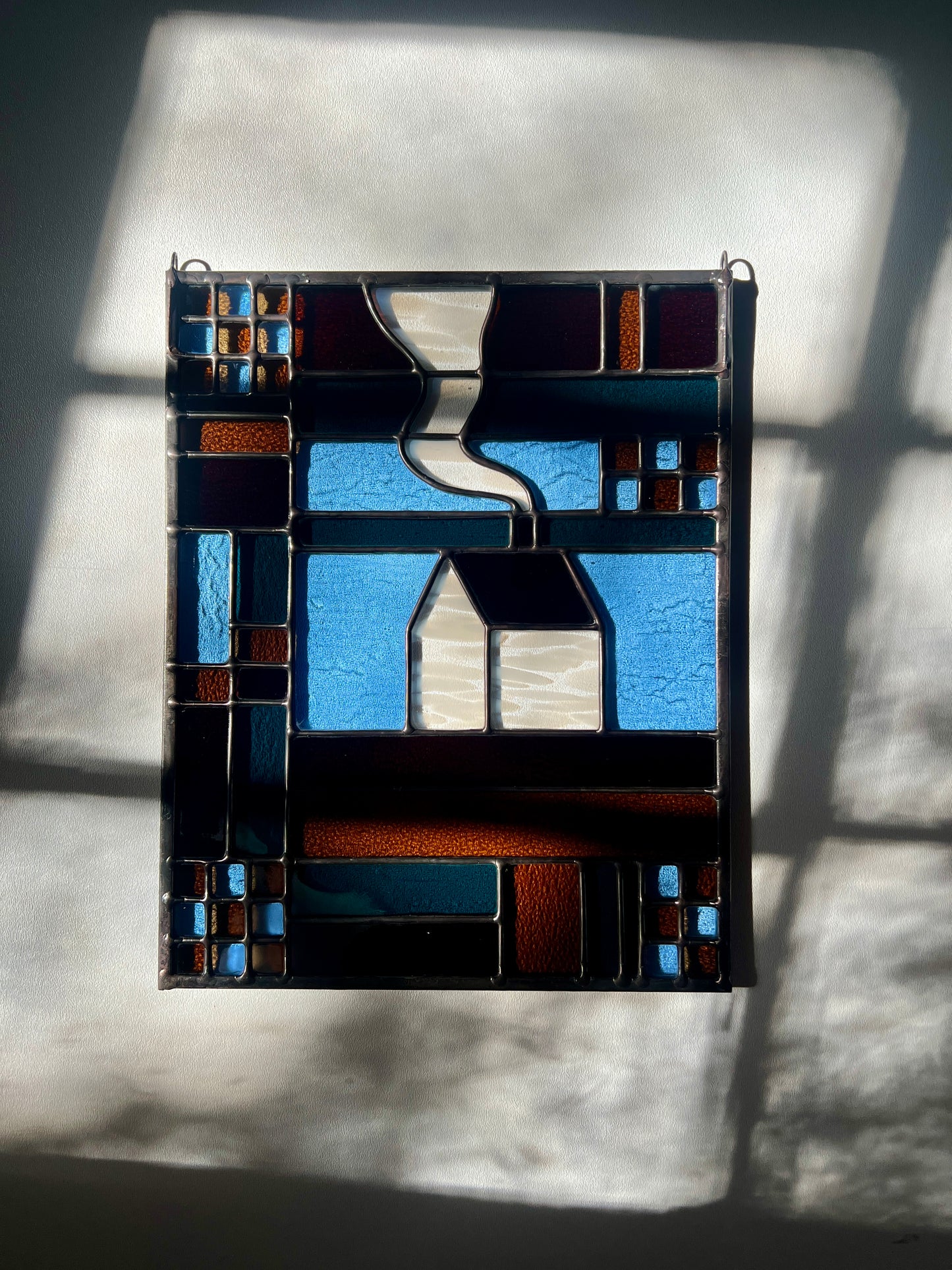 Stained glass textile inspired panel depicting a home in the center in shades of teal, brown, and blue, resting on a white surface with bright sunlight and shadows cast across