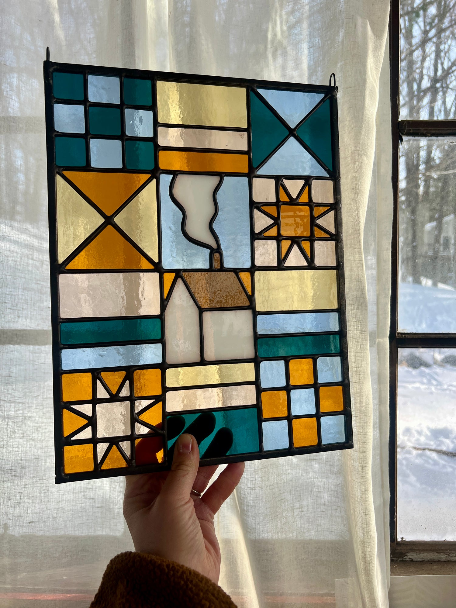 Stained glass textile inspired panel depicting a home in the center in shades of teal, pink, white, blue, and yellow, held by a hand in front of a sheer tan curtain