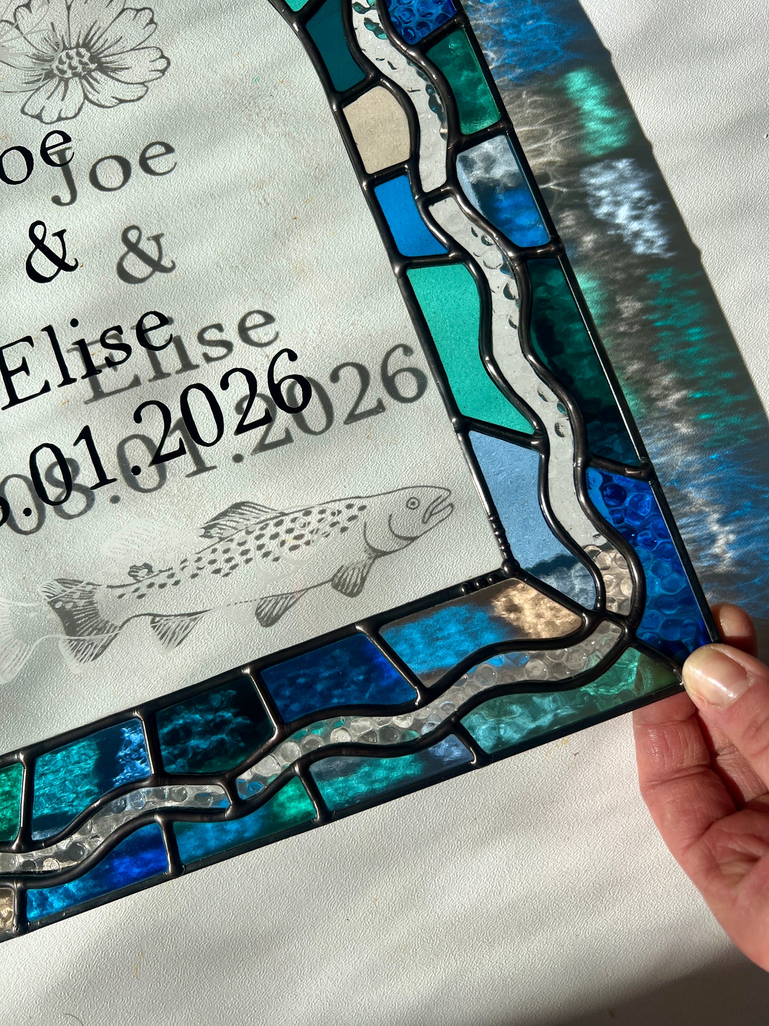 Close up of the corner of a custom stained glass piece, depicting a colorful border in shades of blue and green and custom hand painted text. A glass etching of a trout is casting a shadow in the bright sunlight. The piece is held by a hand.