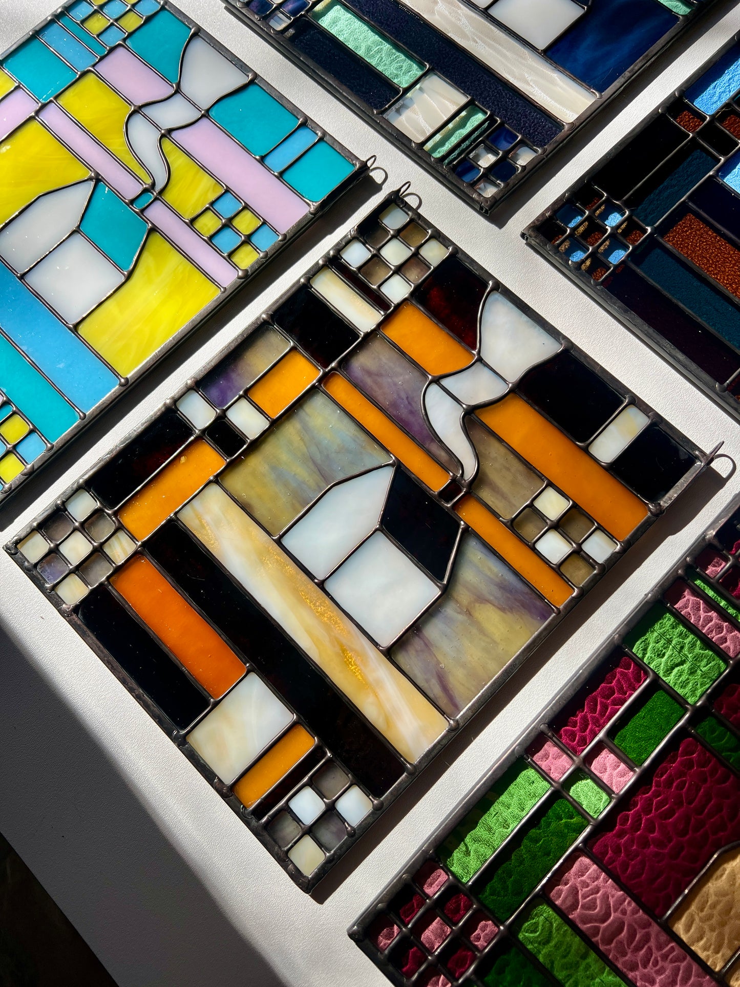 Stained glass textile inspired panel depicting a home in the center in shades of purple, white, brown, and amber,  with panels of the same design surrounding it on a white surface