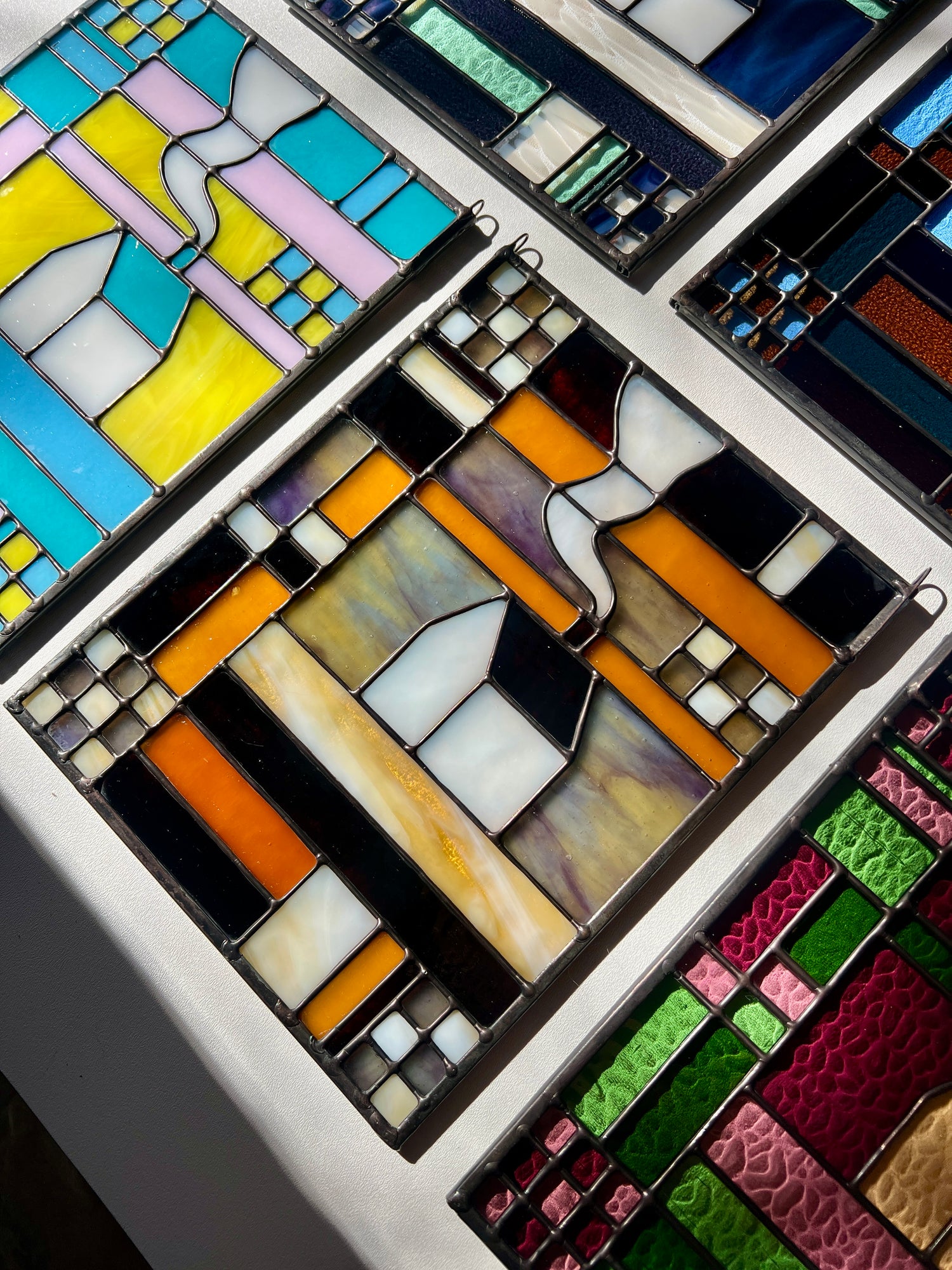 Stained glass textile inspired panel depicting a home in the center in shades of purple, white, brown, and amber,  with panels of the same design surrounding it on a white surface