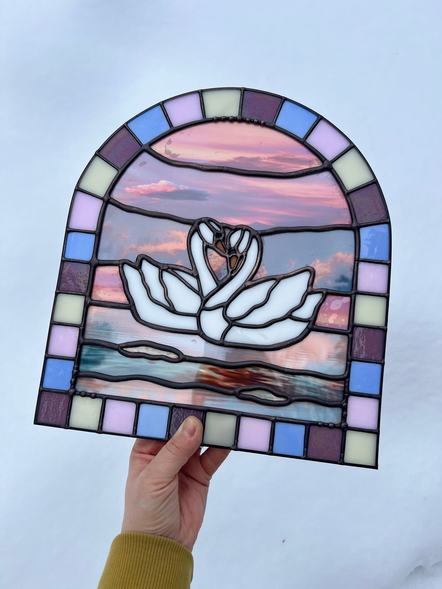 Stained glass panel with a colorful pastel arch border and two white swans in the center. Collaged photographs are used to create a sunset background. The piece is held by a hand against a snowy white background.