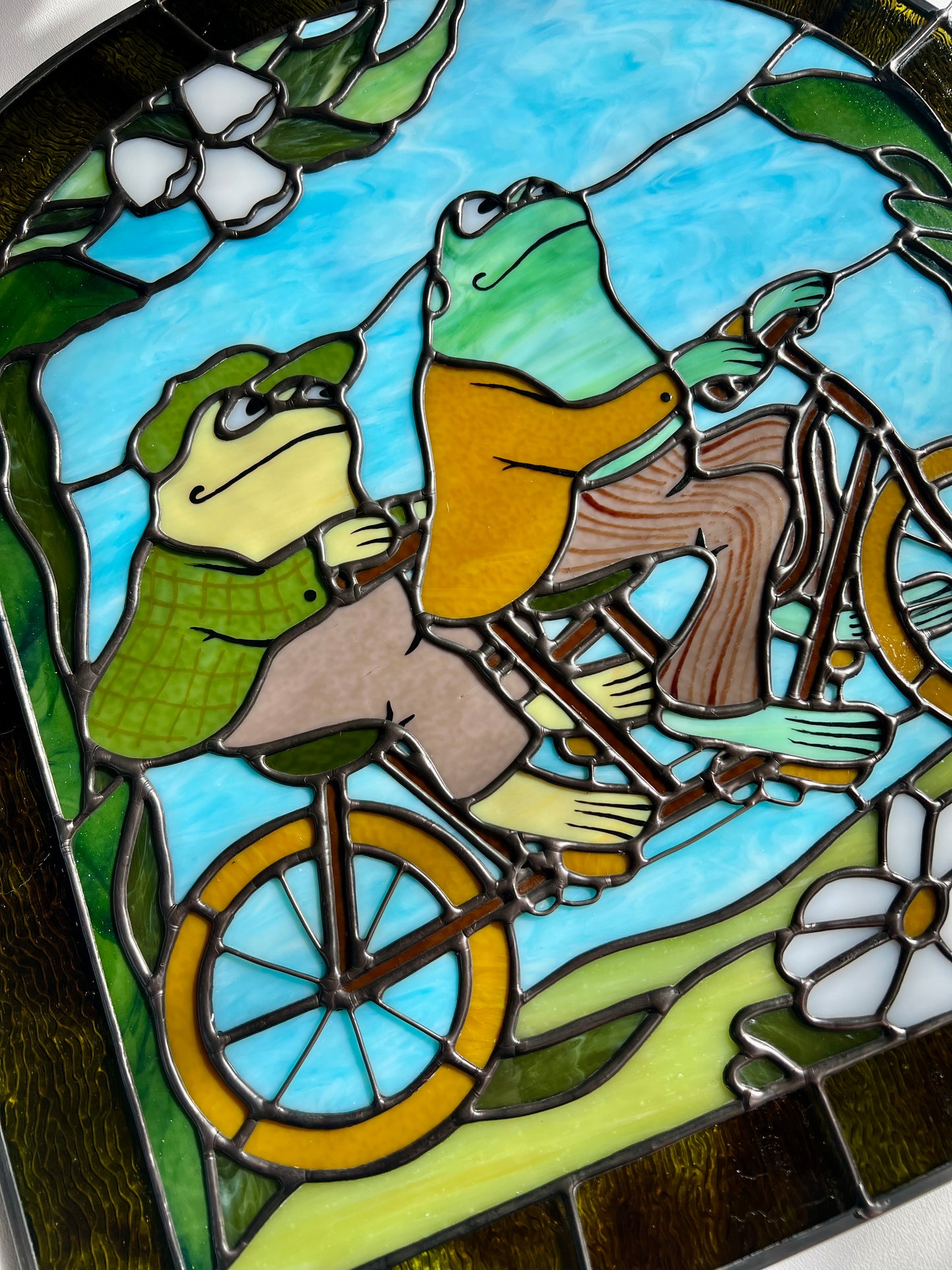close up of a stained glass panel depicting frog and toad riding a bicycle resting on a white surface