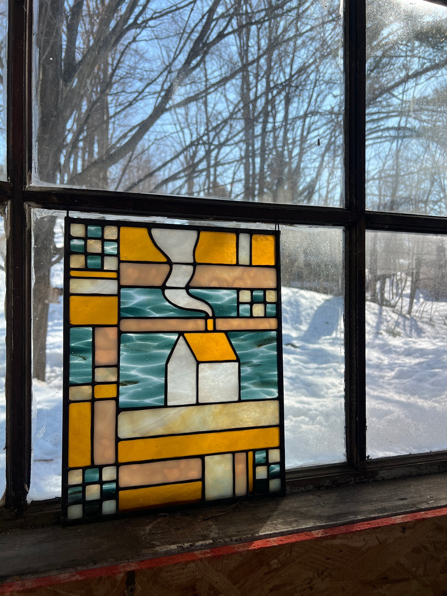 Stained glass textile inspired panel depicting a home in the center in shades of teal, amber, white, and beige, resting against a brightly backlit window with a snowy scene outside