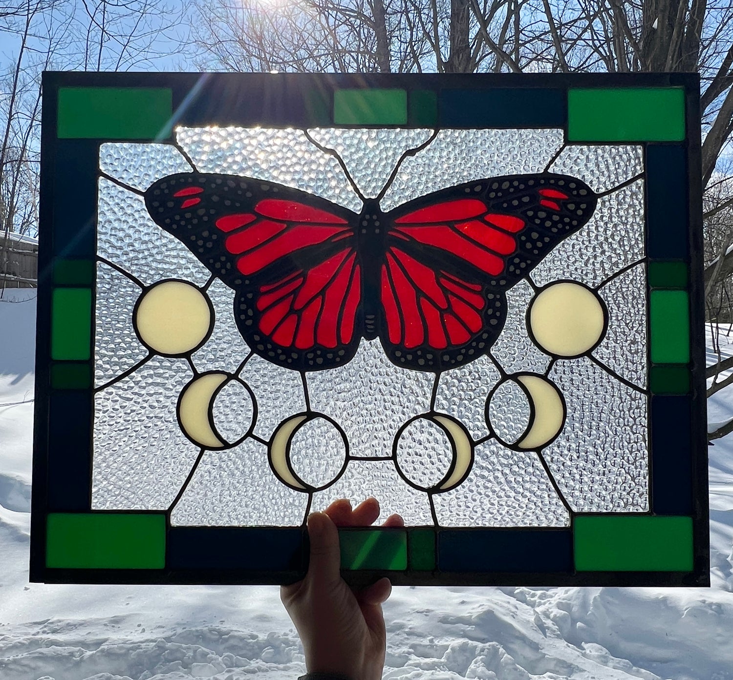 A large stained glass panel with a thick blue and green geometric border and a monarch butterfly and moon phases in the center, with textured clear glass in the background, held by a hand outside against a wintery background with bright sunlight providing backlighting.