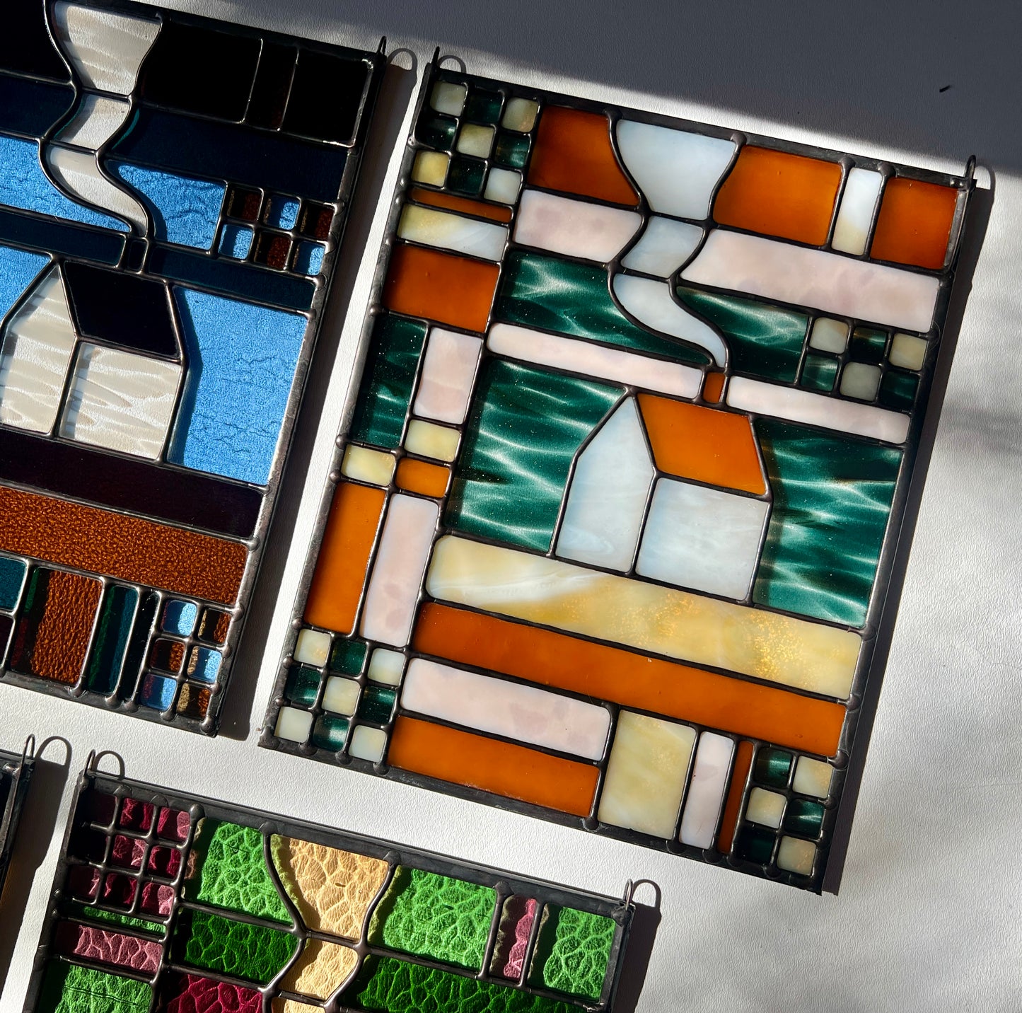 Stained glass textile inspired panel depicting a home in the center in shades of teal, amber, white, and beige, set in a flat lay on a white surface with other similar panels