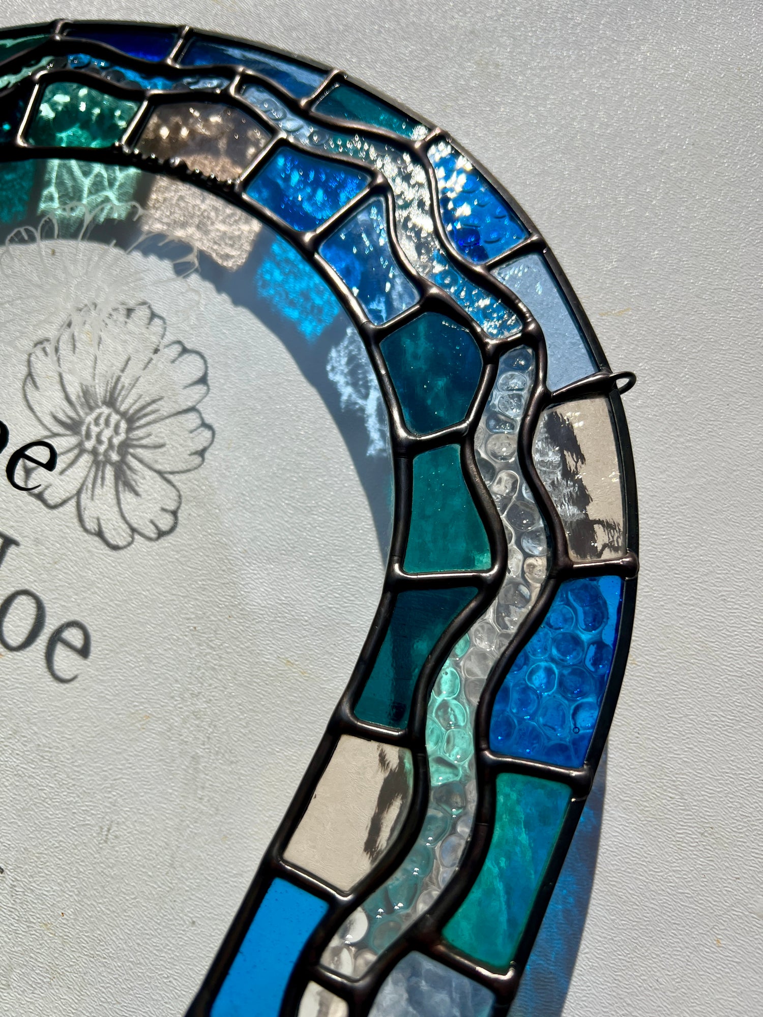 Close up of a stained glass panel in the shape of an arch with a multicolor blue, teal, turquoise, and pink border. Within the border is squiggle detail in clear textured glass. The center of the arch is clear uncolored glass, with personalized text and hand etched details of a cosmos flower. The photograph is taken at an angle to highlight the smooth solder of the border.