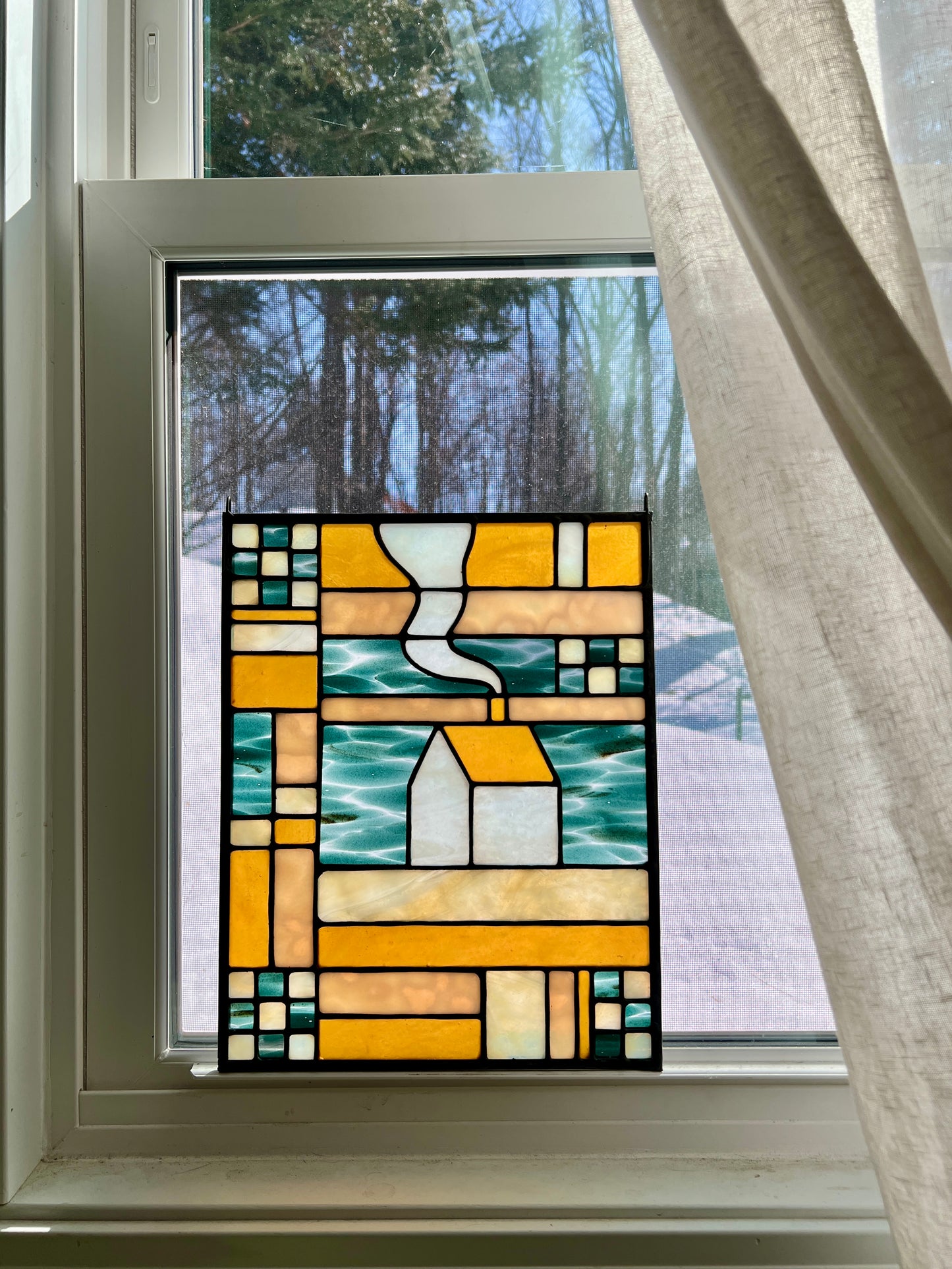 Stained glass textile inspired panel depicting a home in the center in shades of teal, amber, white, and beige, resting in a brightly backlit window with a snowy scene outside