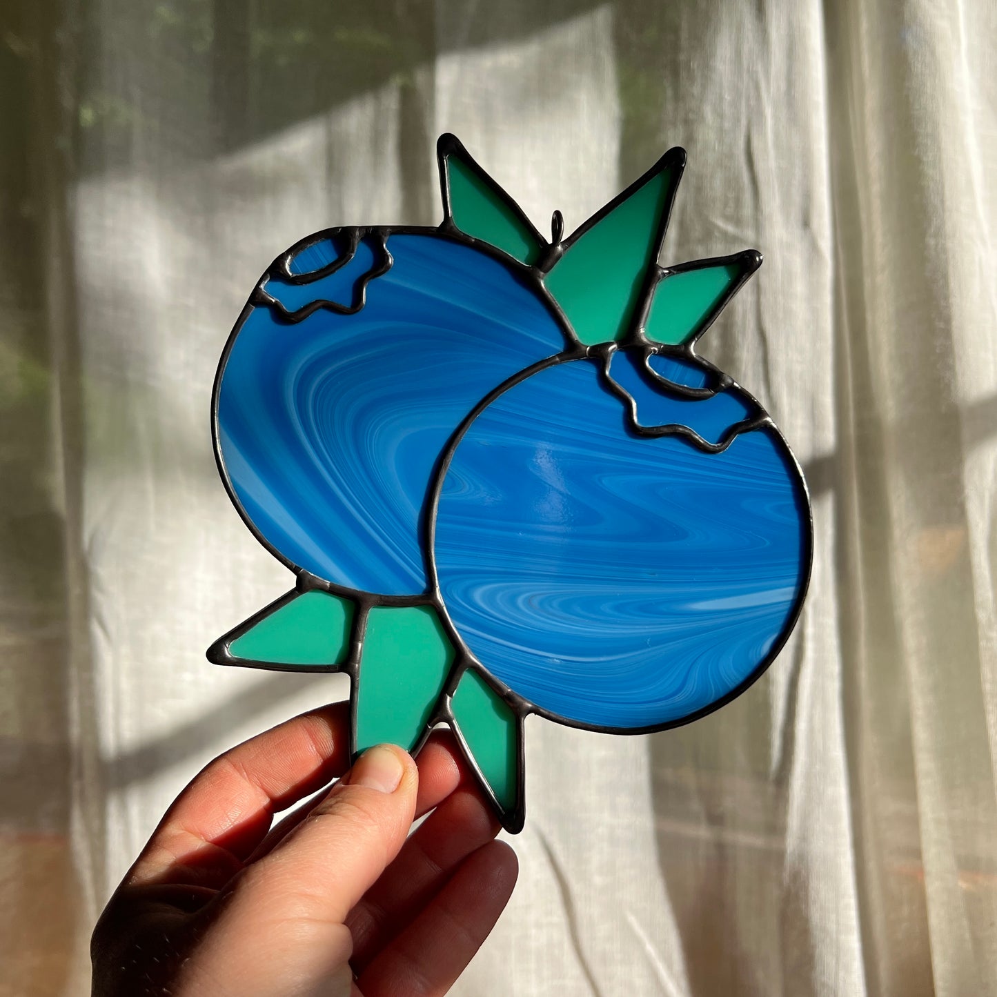 hand holding a streaky blue stained glass suncatcher depicting a small bunch of blueberries with leaves in front of a window