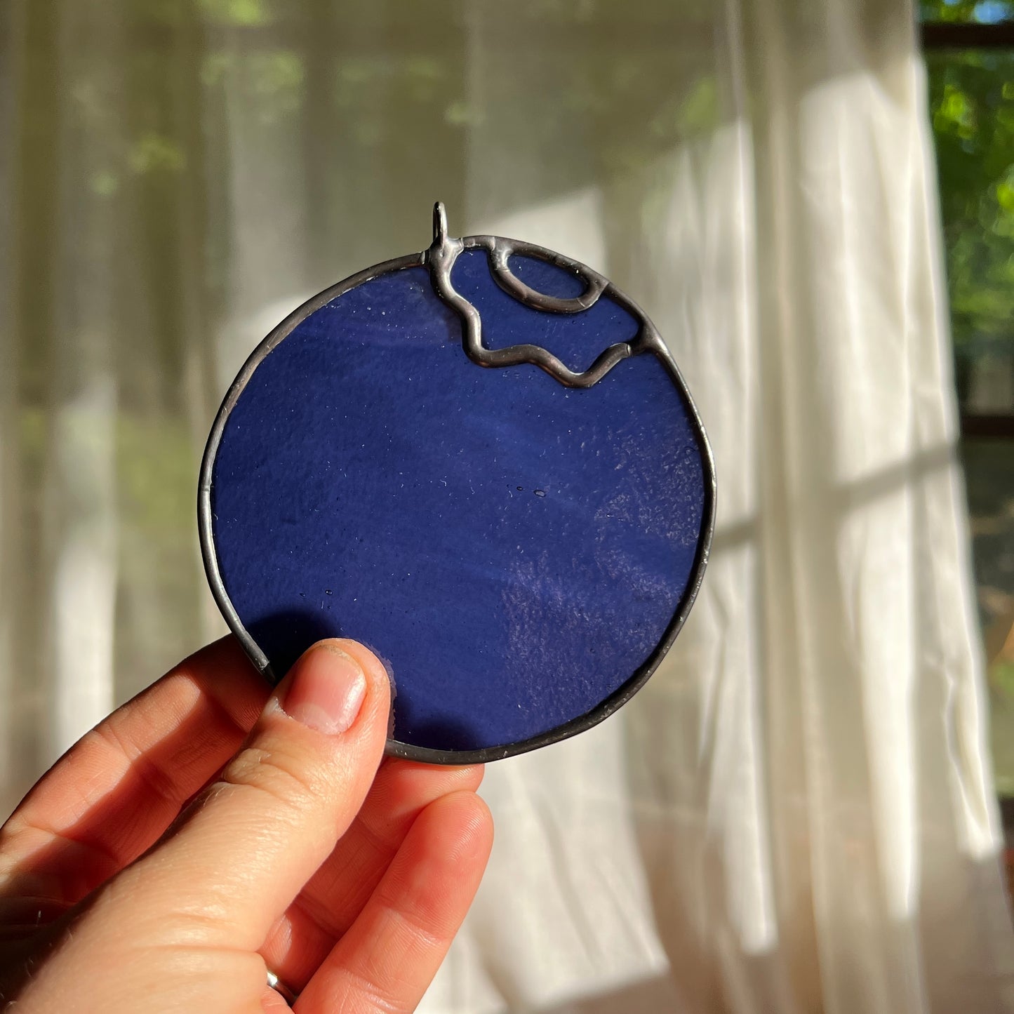 hand holding a wispy blue stained glass blueberry in front of a window