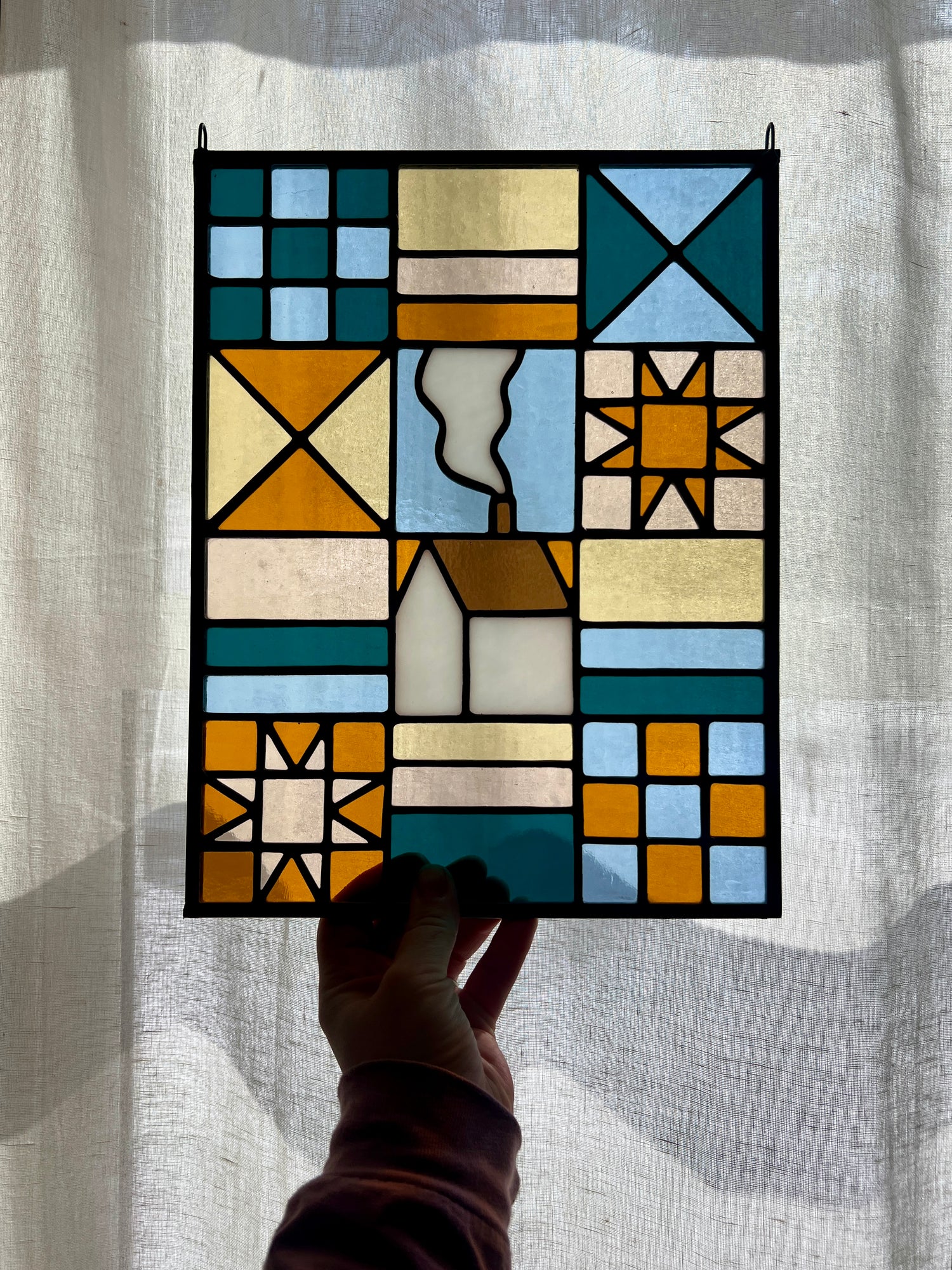 Stained glass textile inspired panel depicting a home in the center in shades of teal, pink, white, blue, and yellow, held by a hand in front of a backlit sheer white curtain