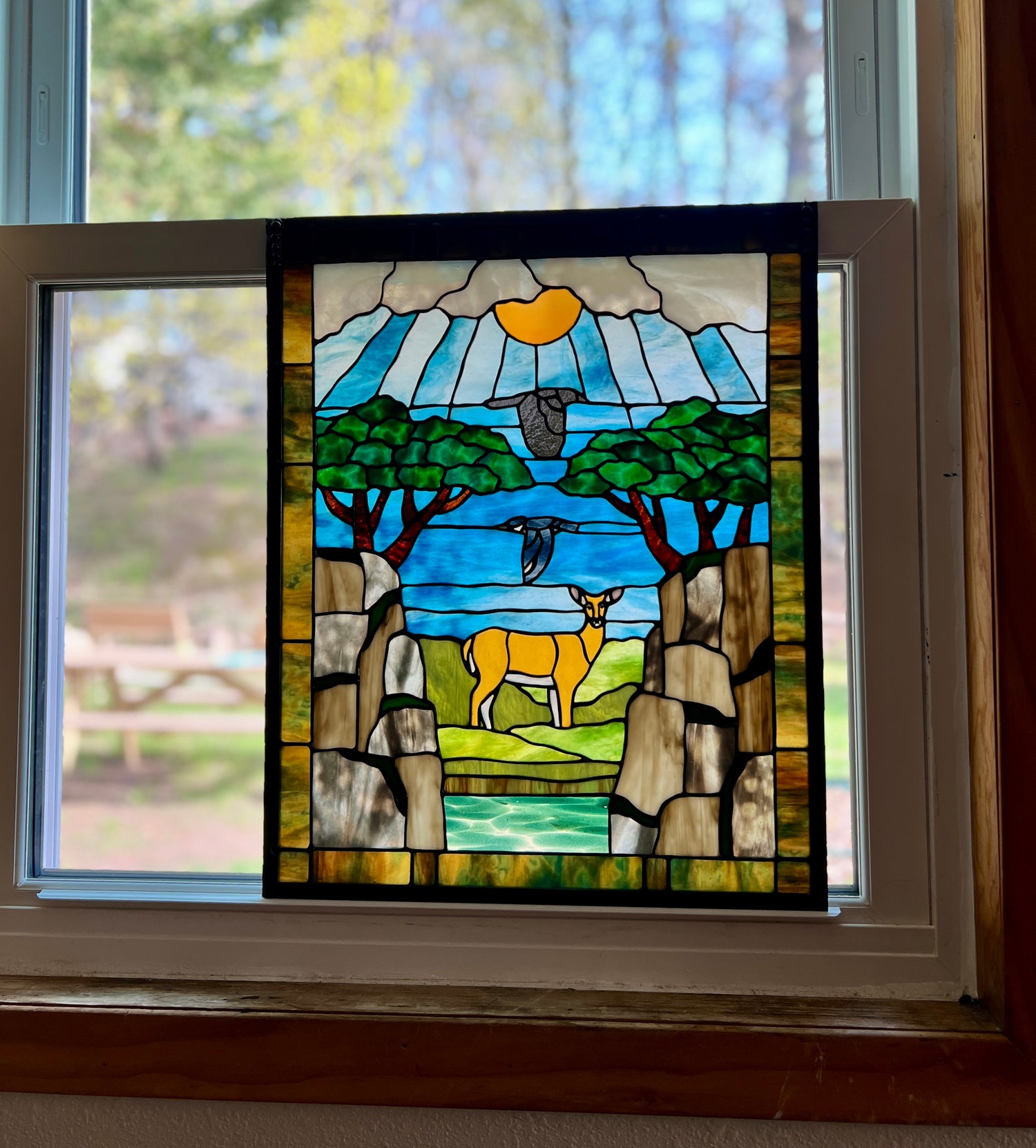 large stained glass panel depicting mossy, rocky cliffs and trees, with a deer, a kingfisher, and a raven standing amongst a river, greenery, and bright blue skies with some clouds and a bright yellow orange sun, resting in a window sill